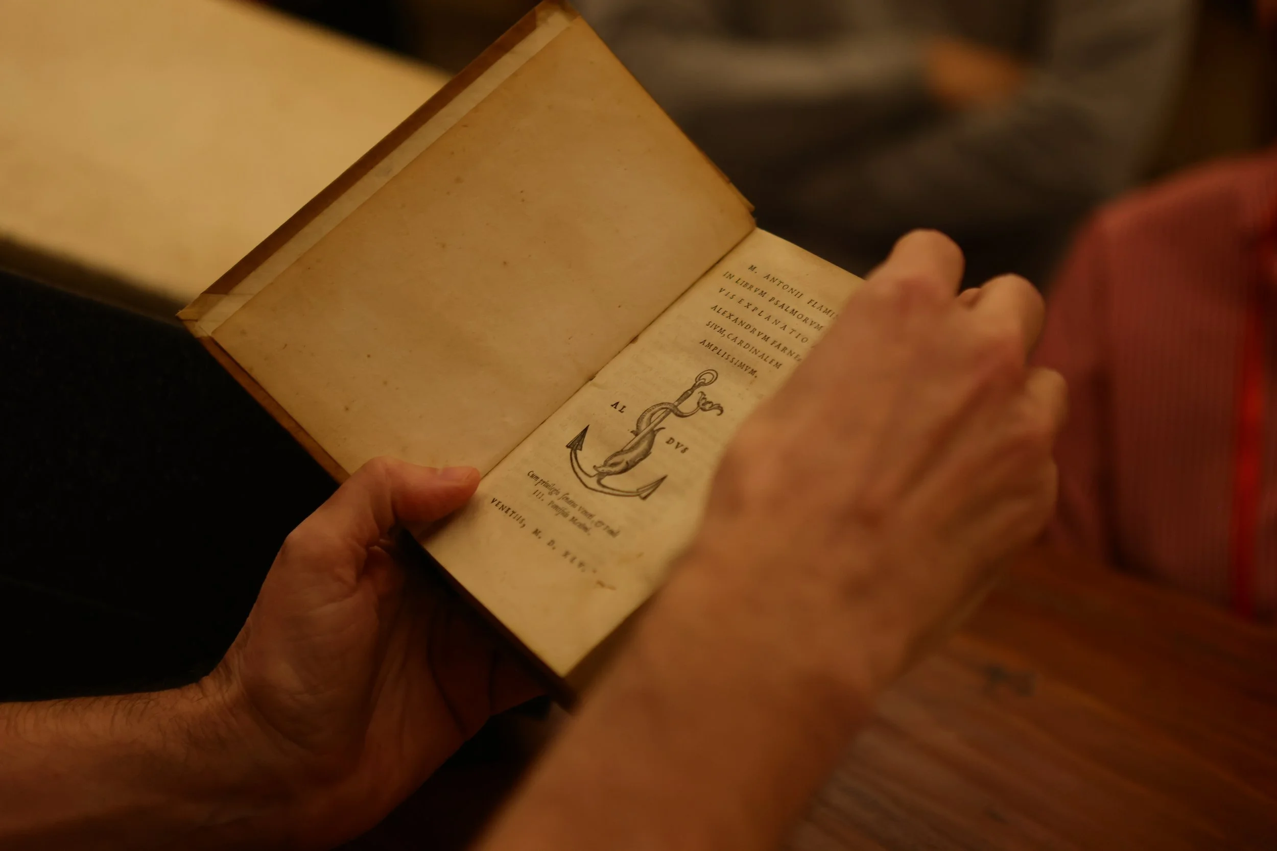 A person holding an old, open book with yellowed pages. The visible page contains Latin text and a sketch of an anchor.