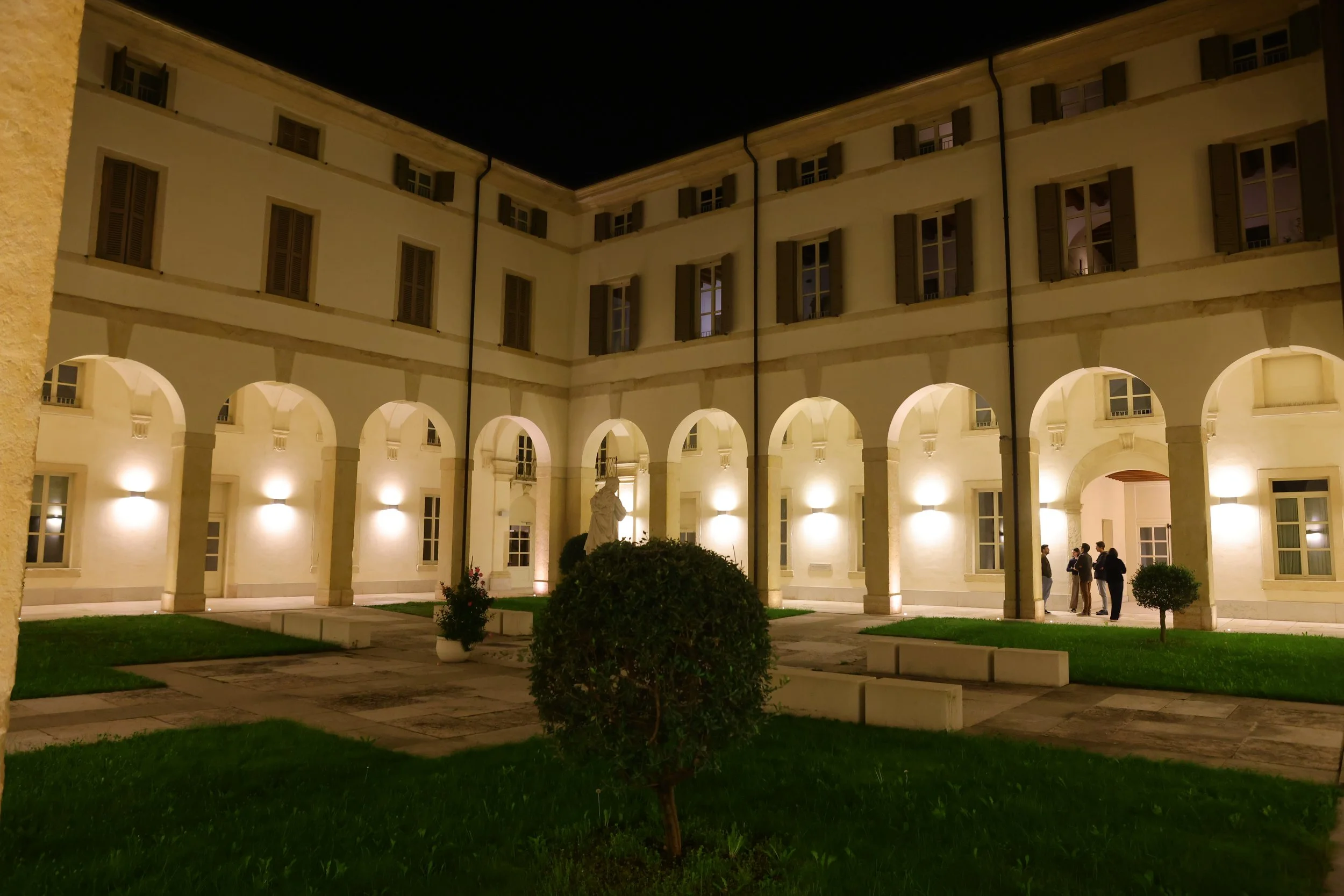 Night view of a courtyard with a cream-colored building featuring arched walkways on the ground level and multiple windows with shutters on the upper levels. The courtyard is illuminated by warm lights, with a statue in the center and a few people standing in conversation.