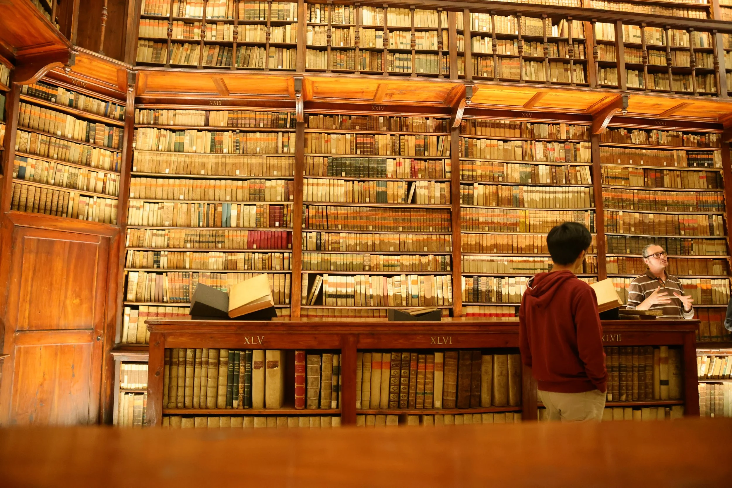 A large wooden bookshelf filled with old, leather-bound books in a library. Two people are standing in front, discussing, with open books in their hands.