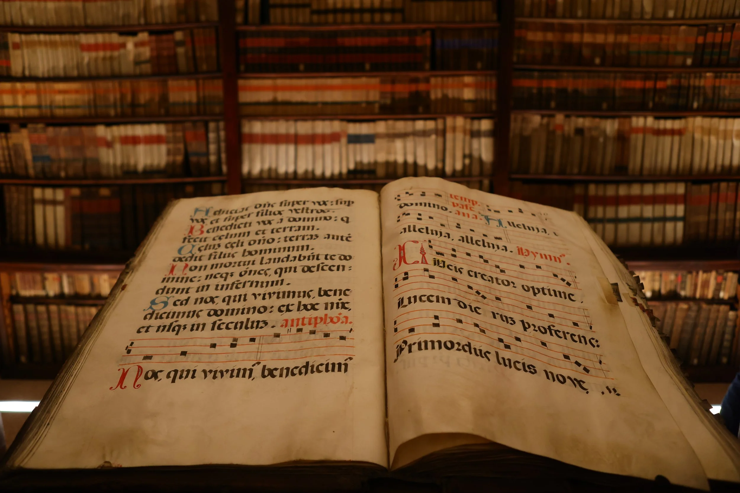 An open medieval illuminated manuscript with Latin text and musical notation, placed in front of a large wooden bookshelf filled with old books.