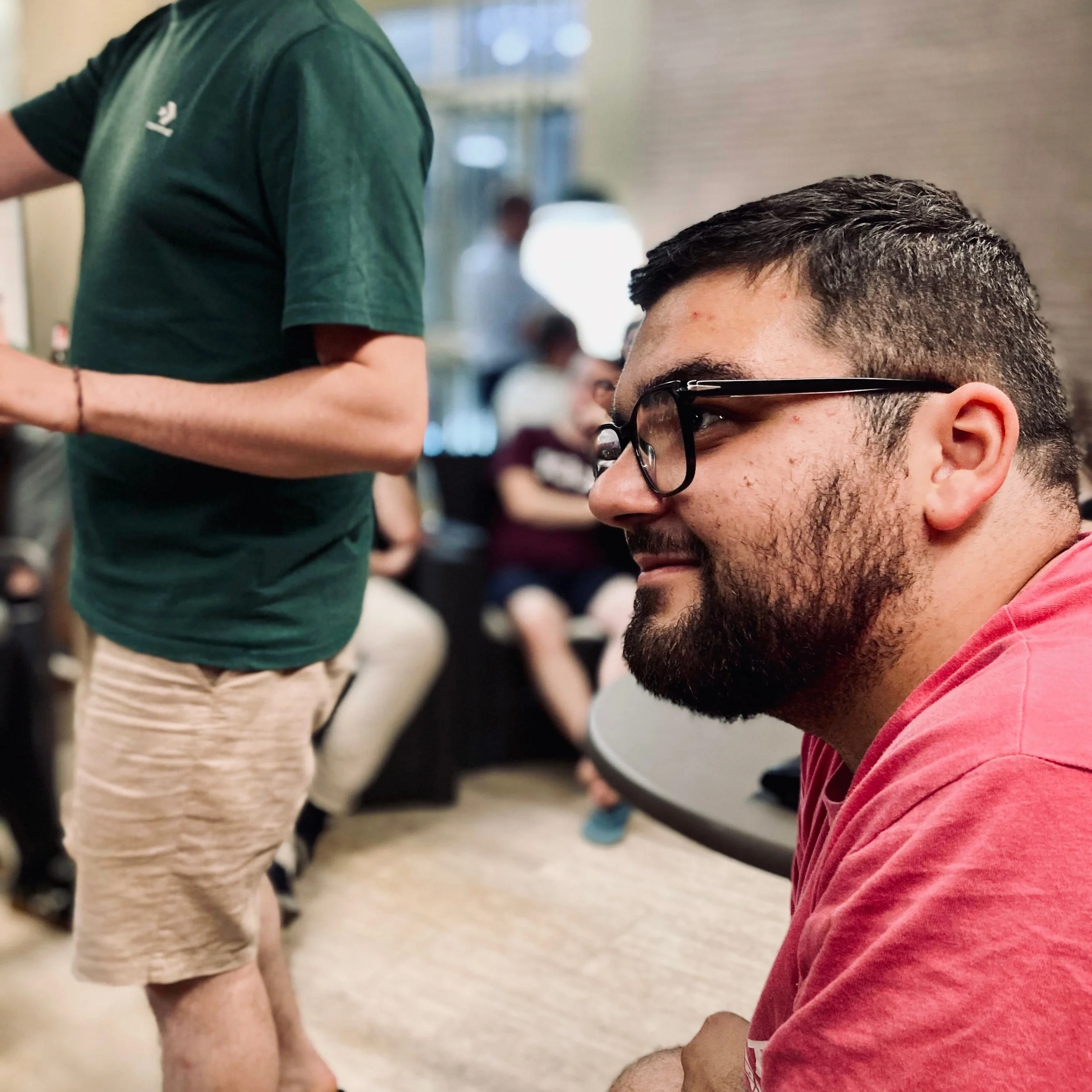 A young man with glasses and a beard is kneeling on the floor while a person in a green shirt and beige pants stands nearby in what appears to be a public indoor space, possibly a waiting area or lobby. There are other people seated in the background.