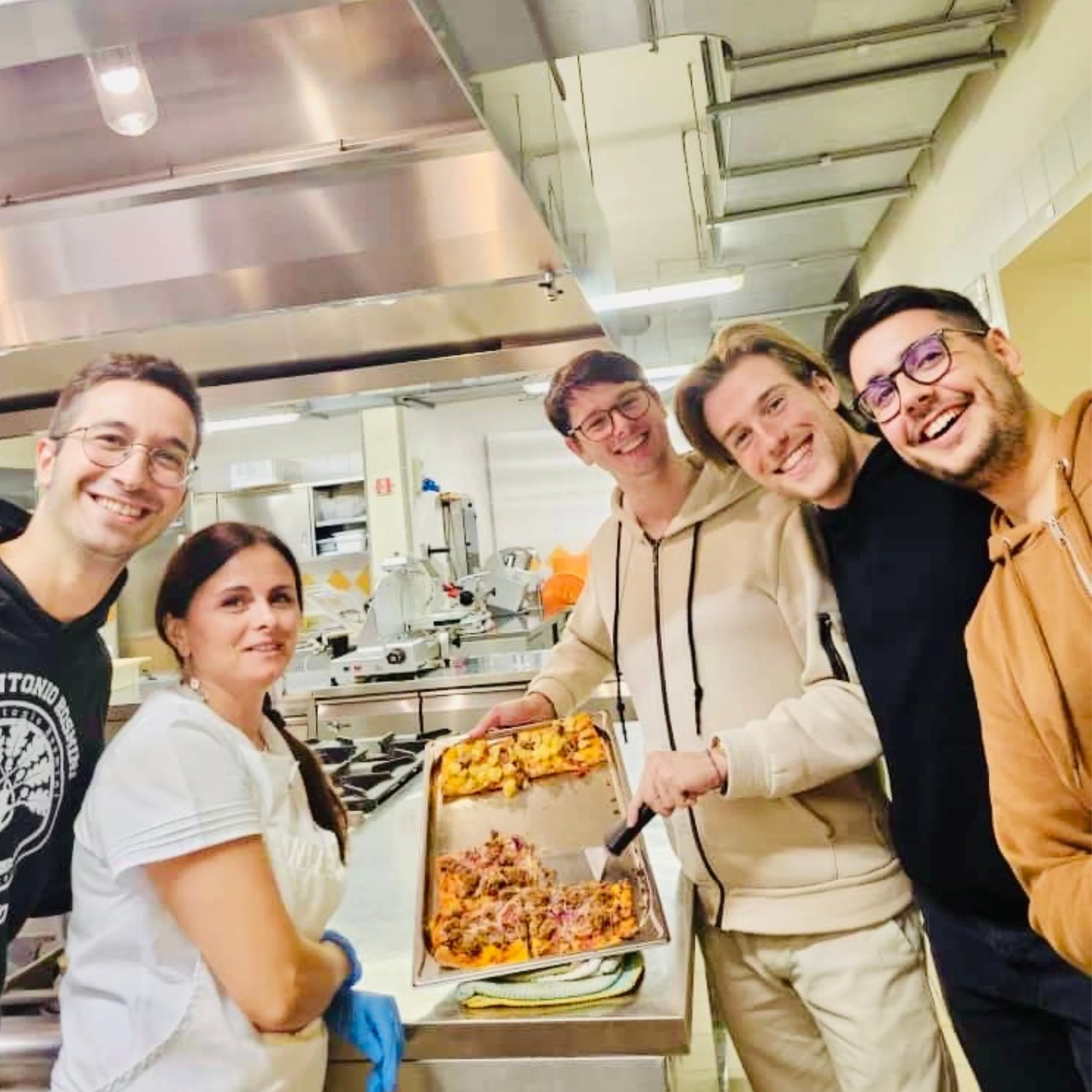 Group of people smiling in a commercial kitchen, holding trays of baked pasta dishes.