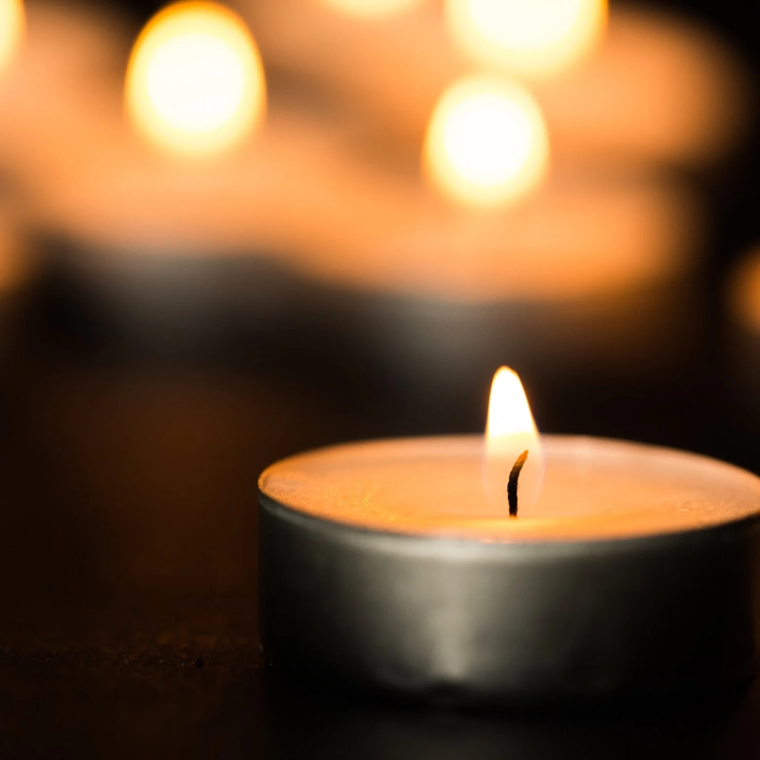 Close-up of a lit tealight candle with a small flame, with blurred warm yellow and orange lights in the background.