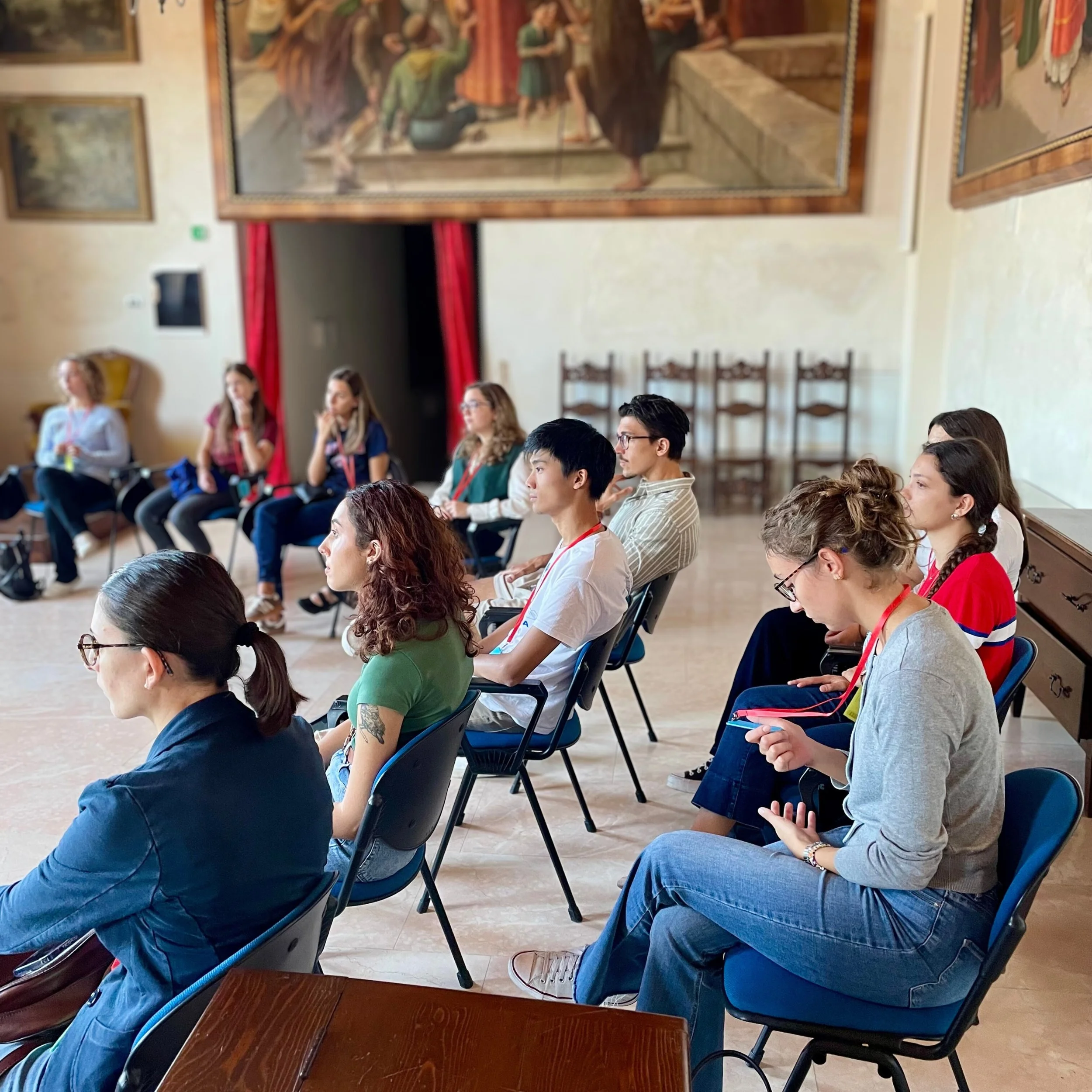People seated in a row, attending an indoor event or conference in a room with religious paintings or murals on the walls.
