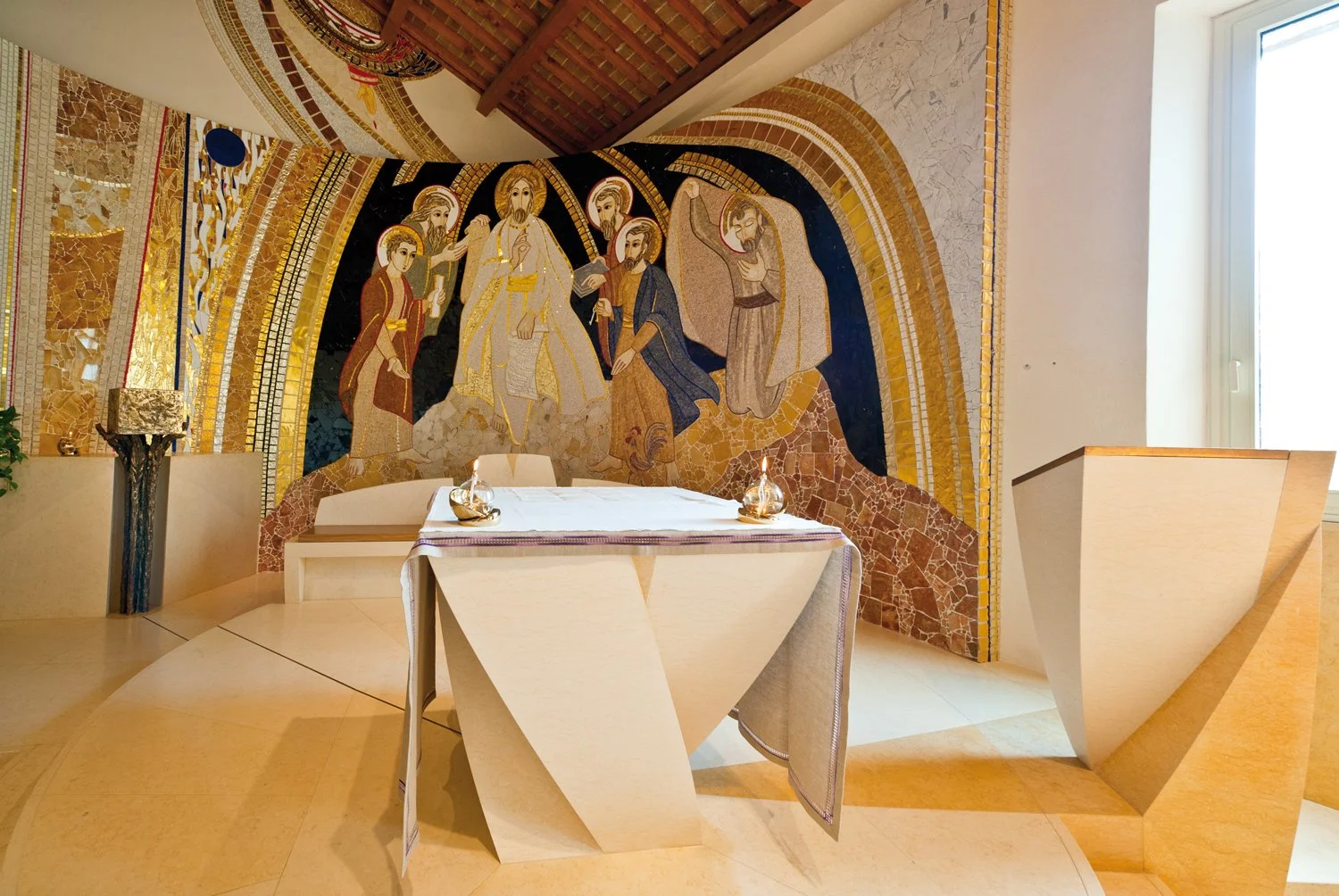 A religious mosaic mural behind an arranged altar with a white tablecloth, candle holders, and decorative items, in a room with a large window to the right.