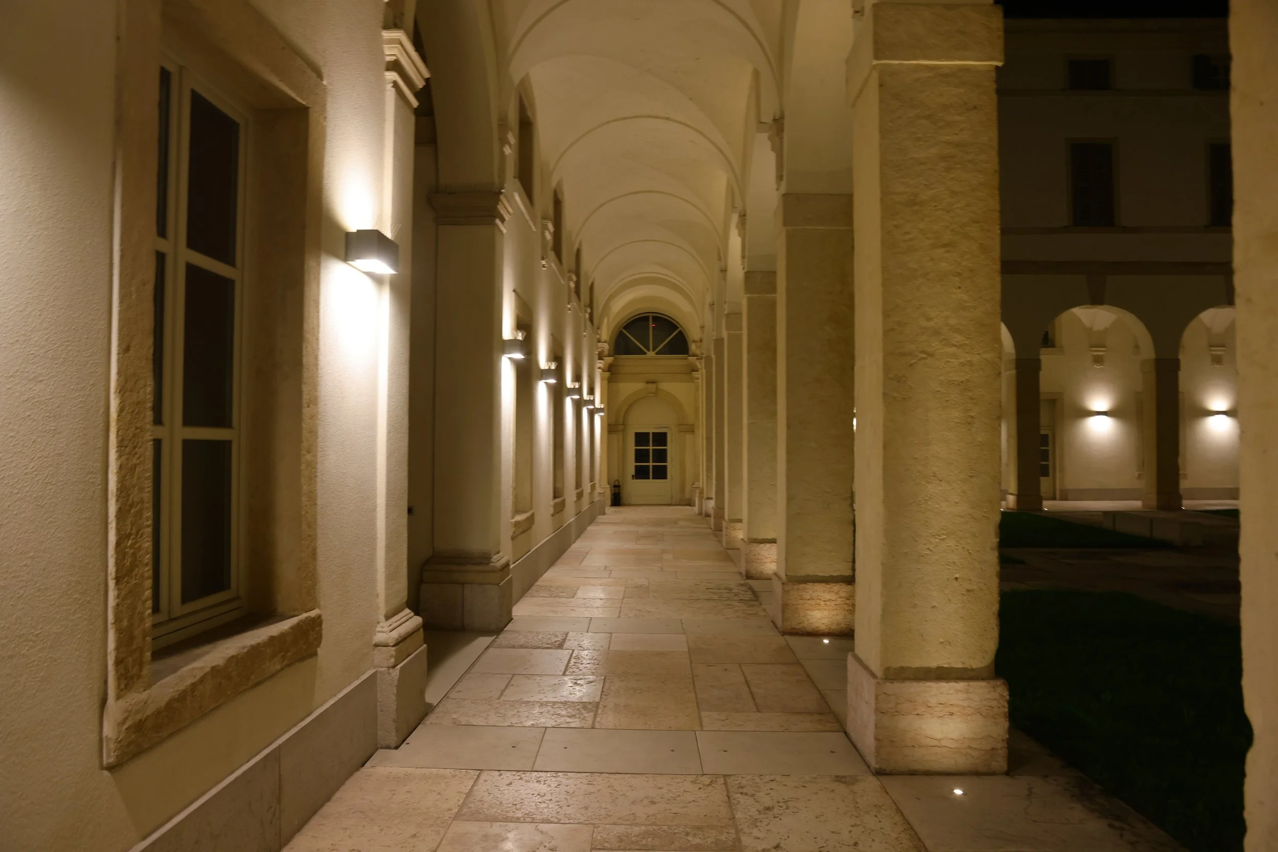 Night view of a covered outdoor corridor with beige stone columns, arches, and walls illuminated by wall sconces, leading to a closed door at the end.