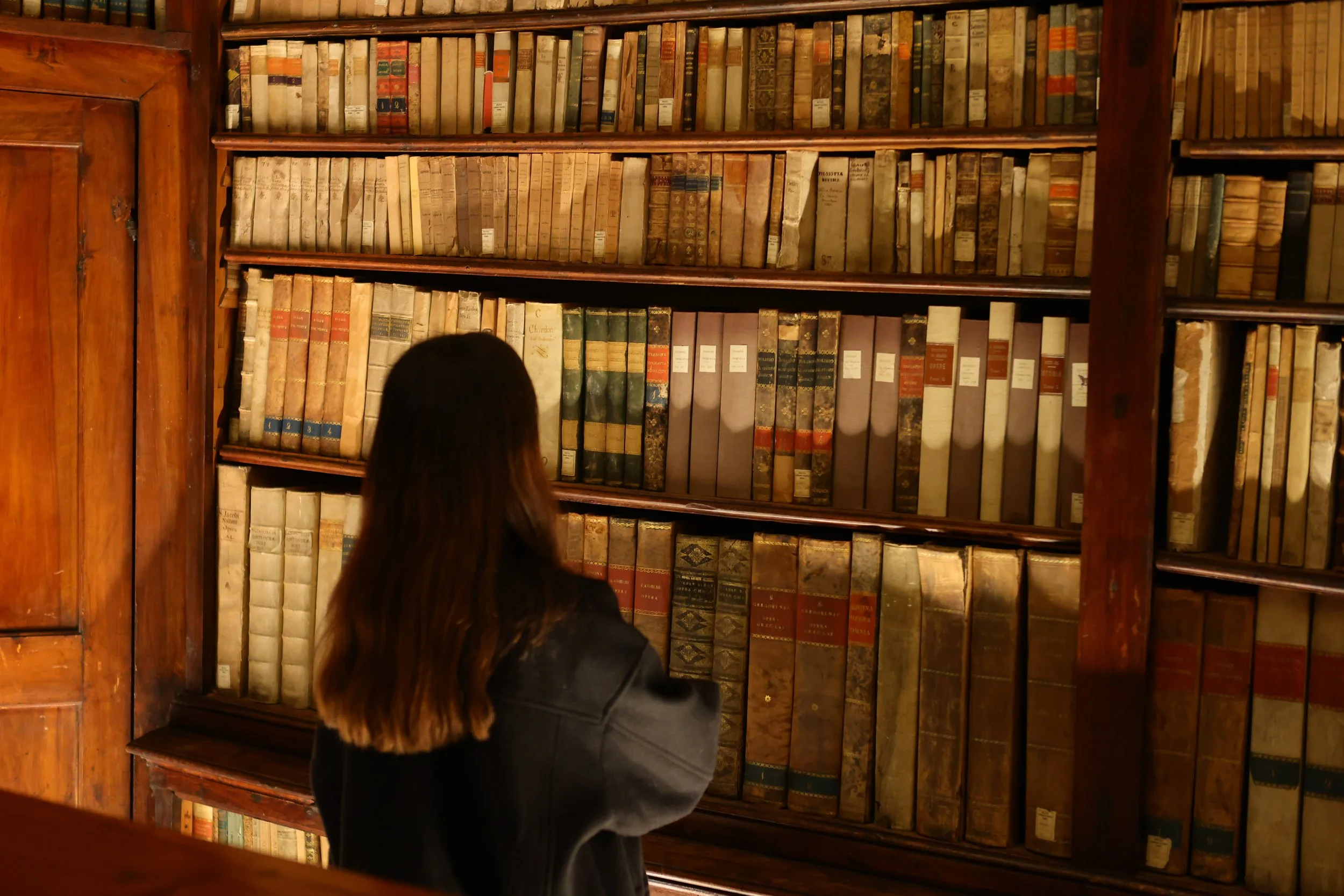 Person with long hair viewing old books on wooden shelves in a library.