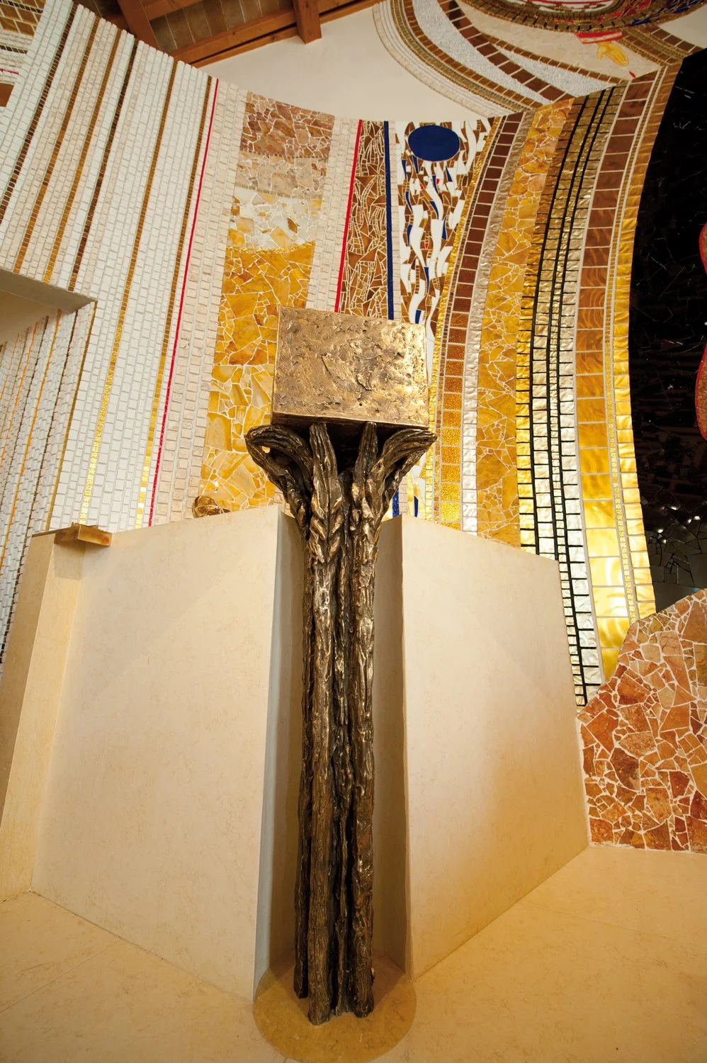 Close-up view of a bust sculpture of a human head on a decorative pedestal with a powerful, textured building interior featuring multicolored tile and mosaic designs in the background.