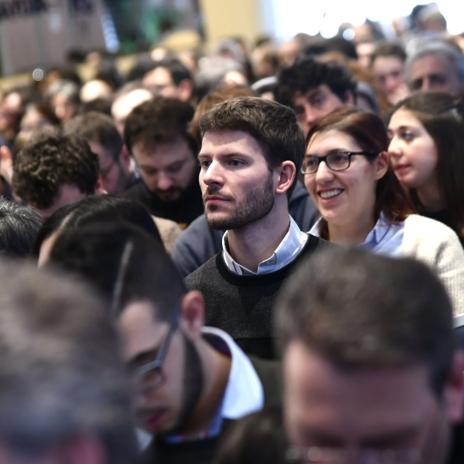 A crowd of people at an indoor event, with a young man with a beard and short hair in the center, and a woman with glasses smiling beside him.