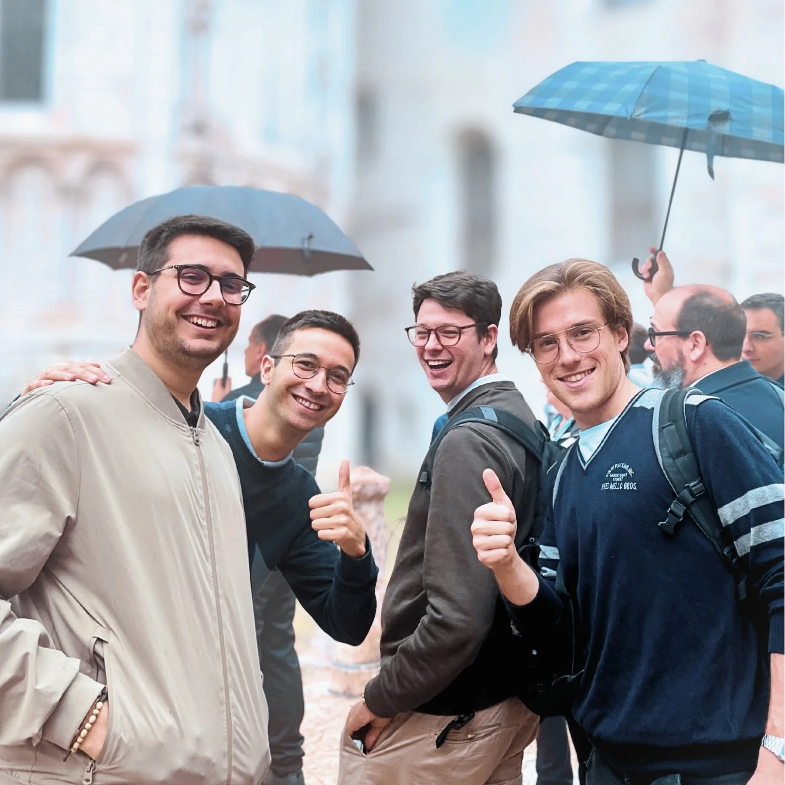 Group of six smiling men with backpacks and glasses, some giving thumbs up, standing close together outdoors on a rainy day, with some holding umbrellas, in front of historic buildings.