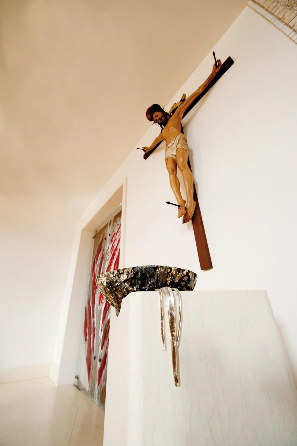 Religious crucifix of Jesus Christ mounted on a white wall with a decorative bowl and a glass hanging from a ledge in the foreground.