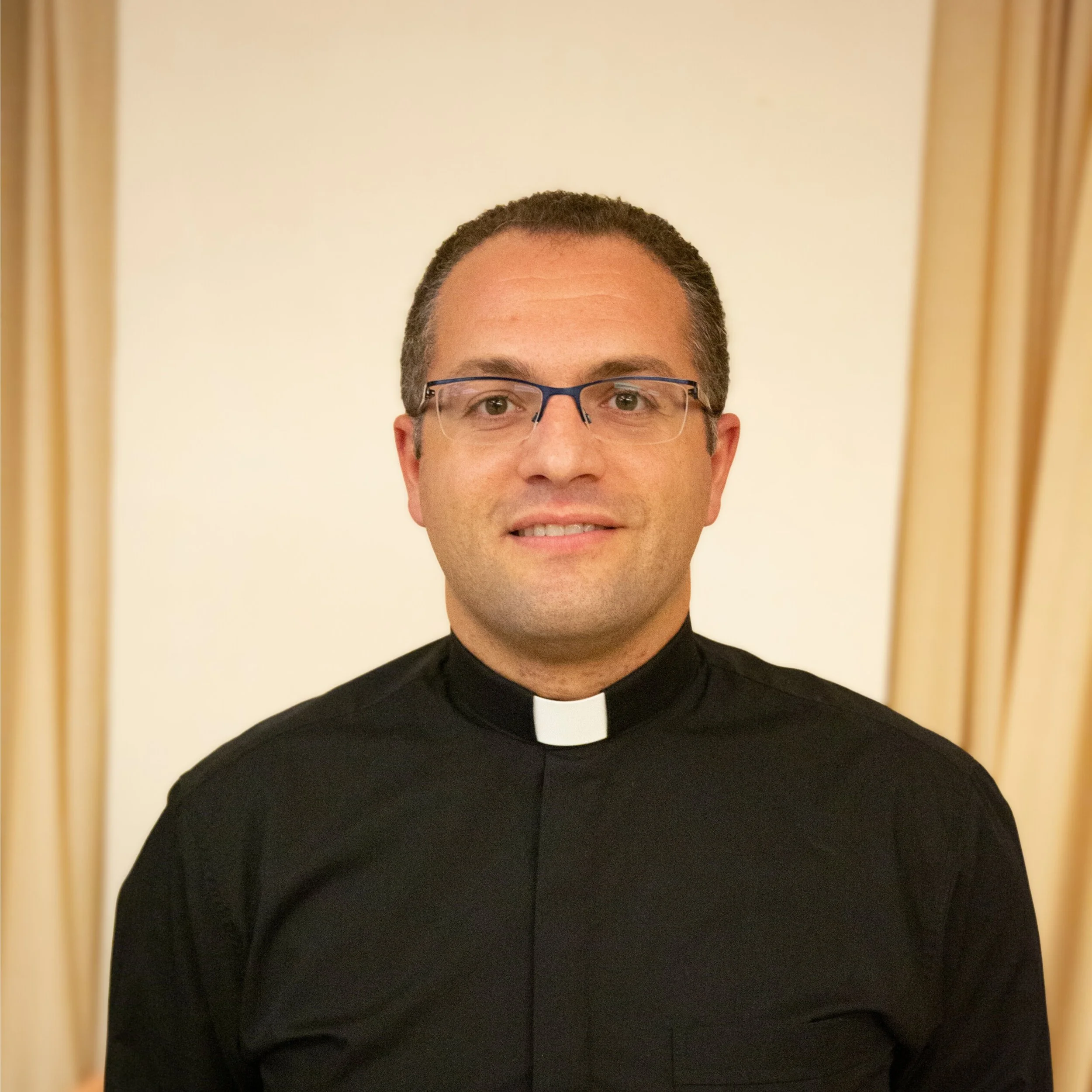 Close-up of a person wearing glasses and a black clergy shirt with a white clerical collar, standing in front of a mirror with a beige frame.