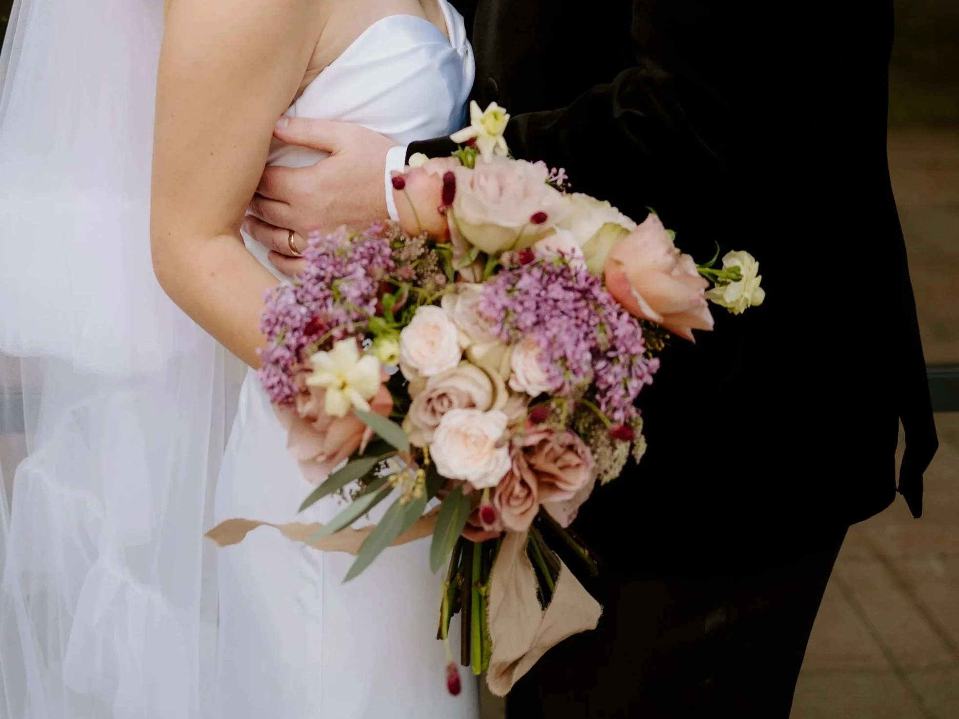 Close-up of a bride holding a bouquet of pink, white, and purple flowers, with a groom wearing a black suit beside her.