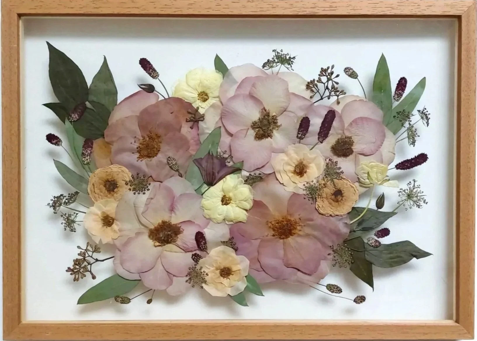 Pressed dried flowers arranged in a framed collage, featuring pink, yellow, and cream-colored blossoms with green leaves and small purple accents.