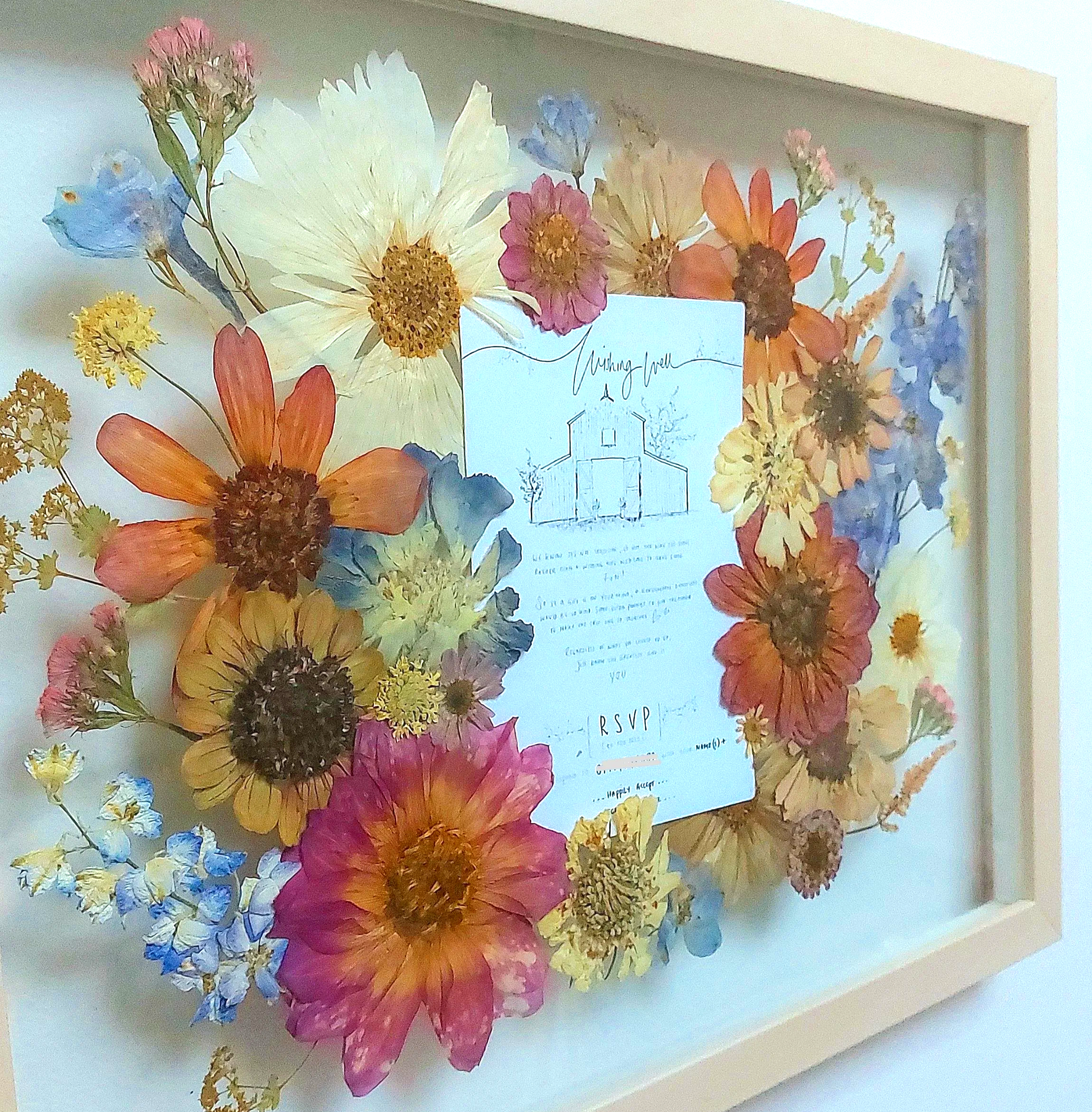 Dried and pressed colorful flowers arranged around wedding invitation in a shadow box frame.