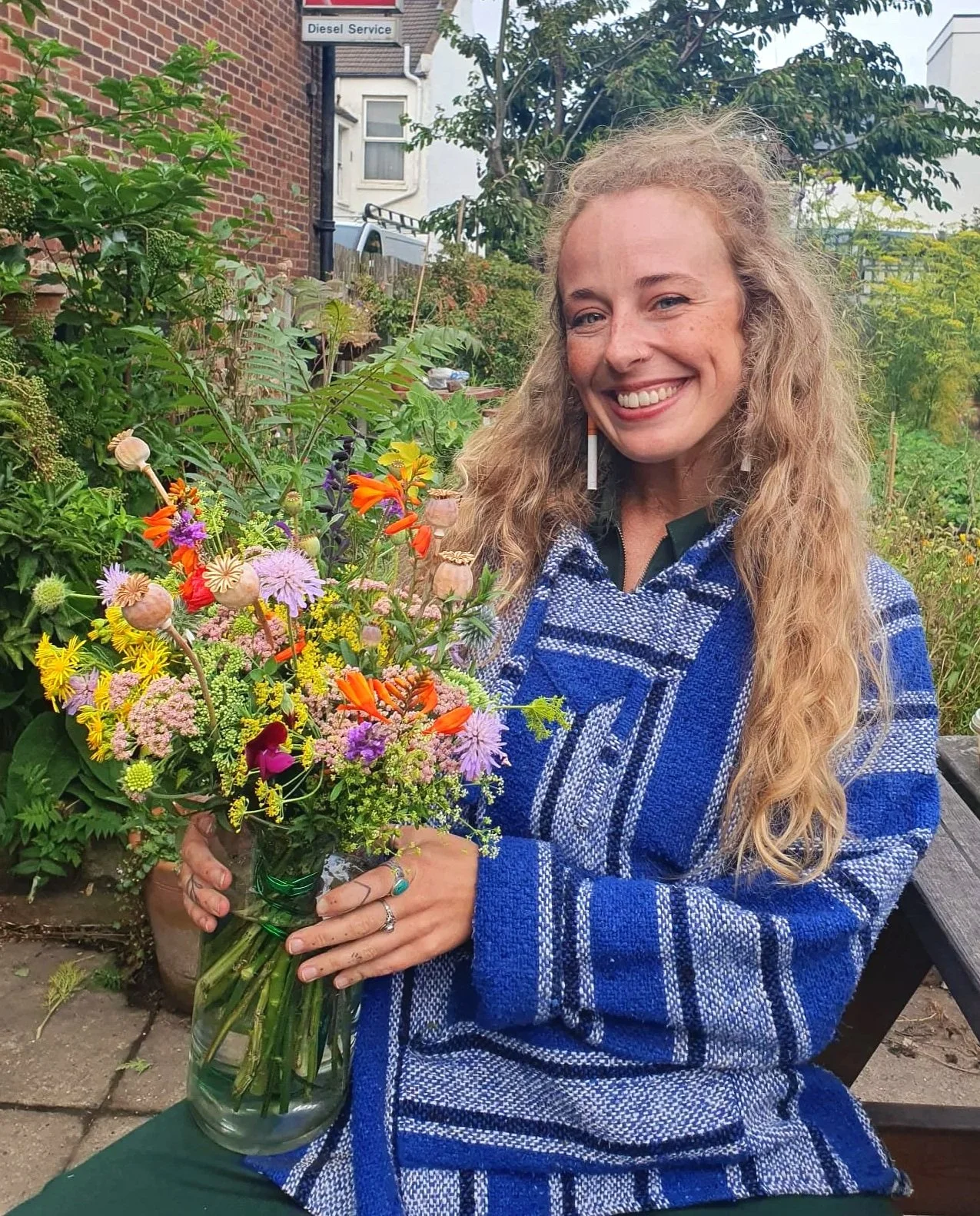 A woman with long curly blond hair smiling and holding a large bouquet of colorful flowers in a glass vase in an outdoor garden setting.