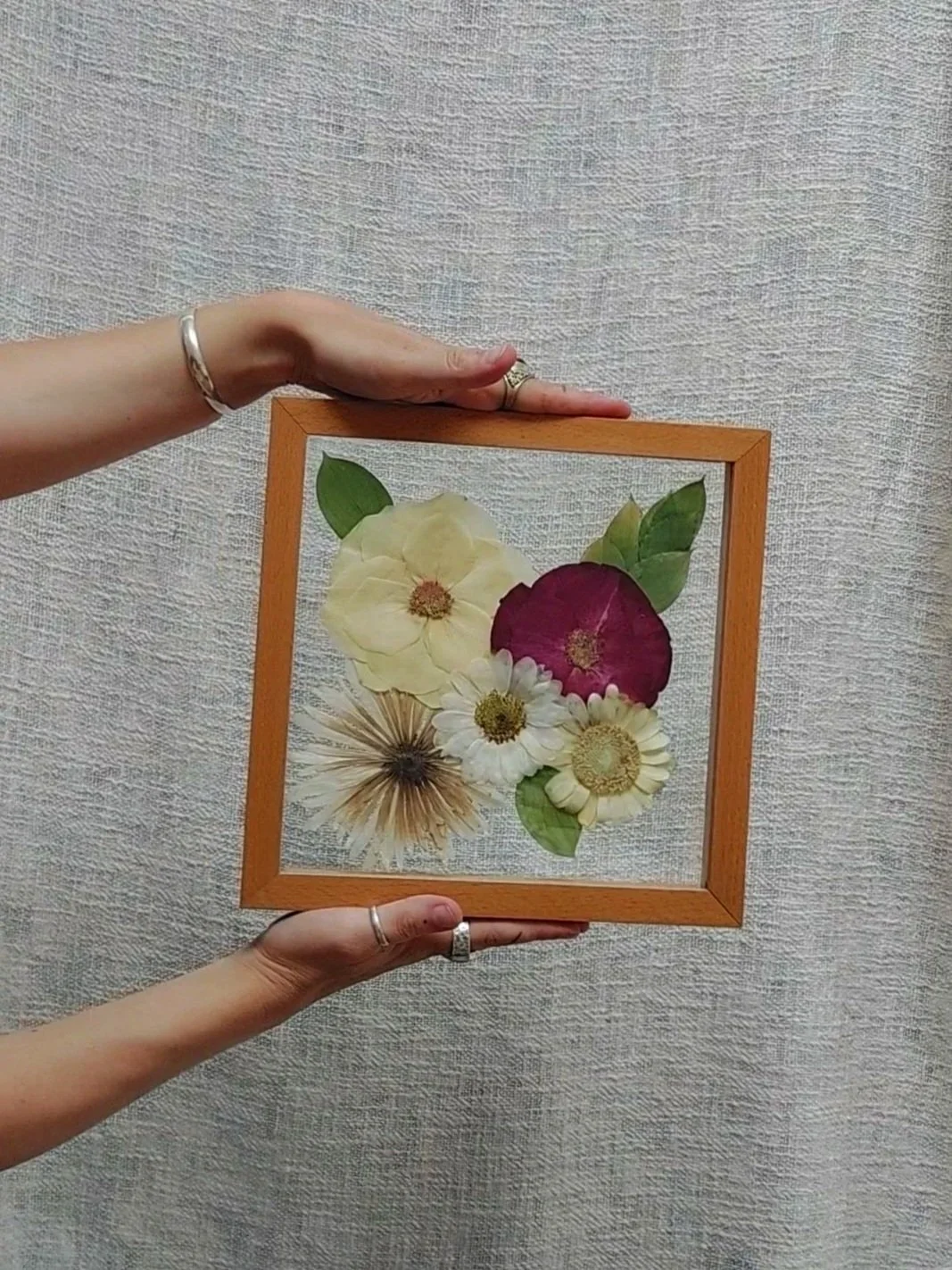 Hands holding a wooden frame containing pressed flowers and green leaves against a textured background.