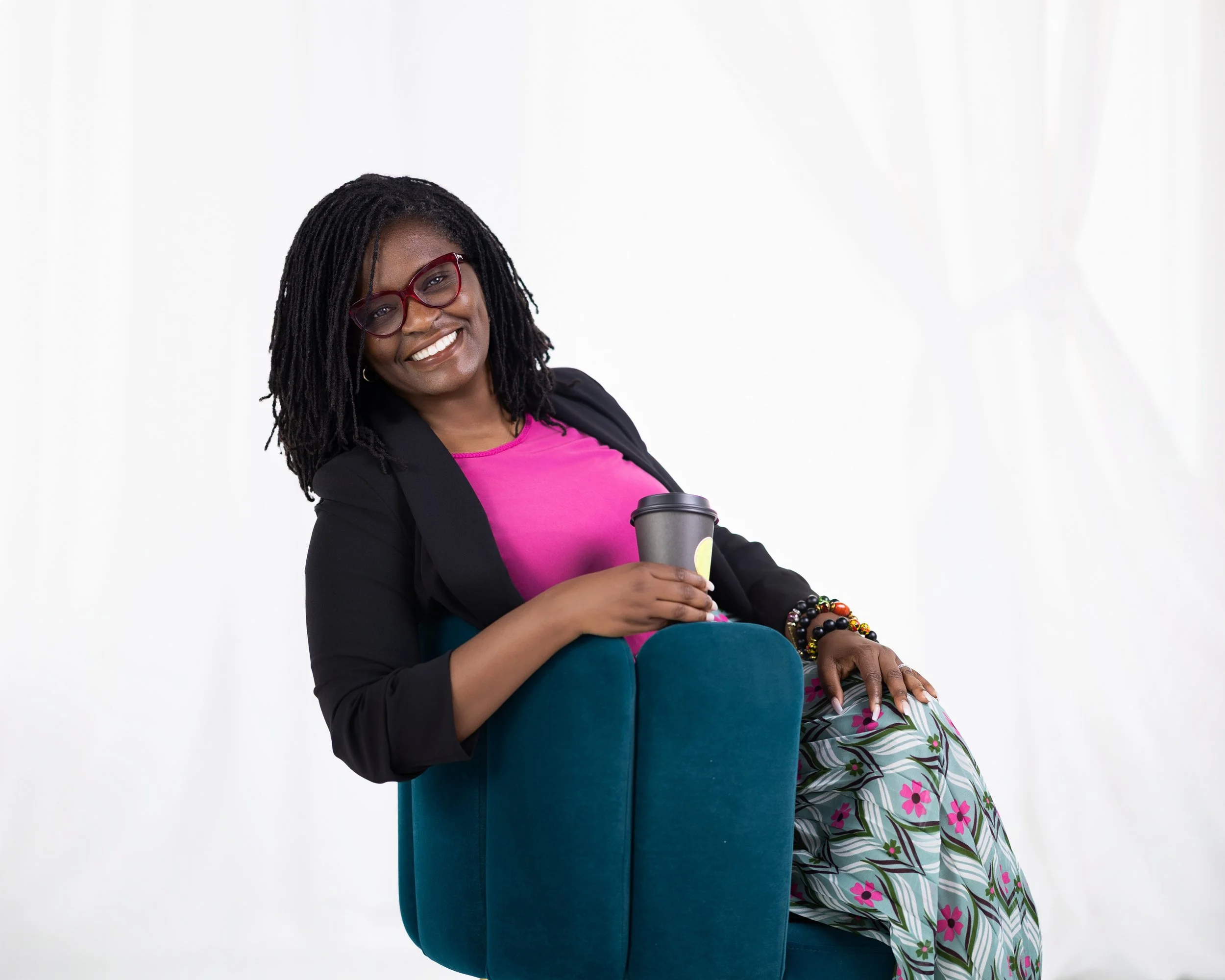 A woman with dark skin, glasses, dreadlocks, and a cheerful smile is sitting on a blue chair against a white background. She is wearing a black blazer over a pink top, decorated with colorful beaded bracelets, and holding a coffee cup.