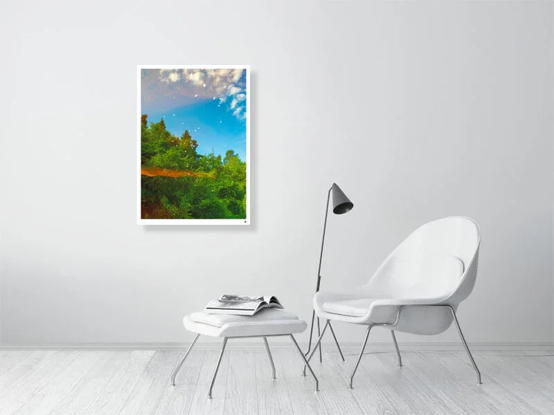 Minimalist room with white chair, small ottoman with open book, black floor lamp, and framed landscape photograph of a sunny sky over green trees on a white wall.