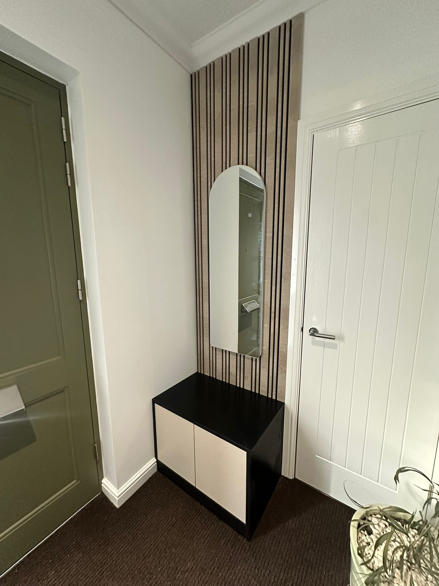 A modern foyer area with a black and white cabinet, a tall mirror on a wood-paneled wall, a white door, and a green door to the left. There is also a potted plant on the floor.