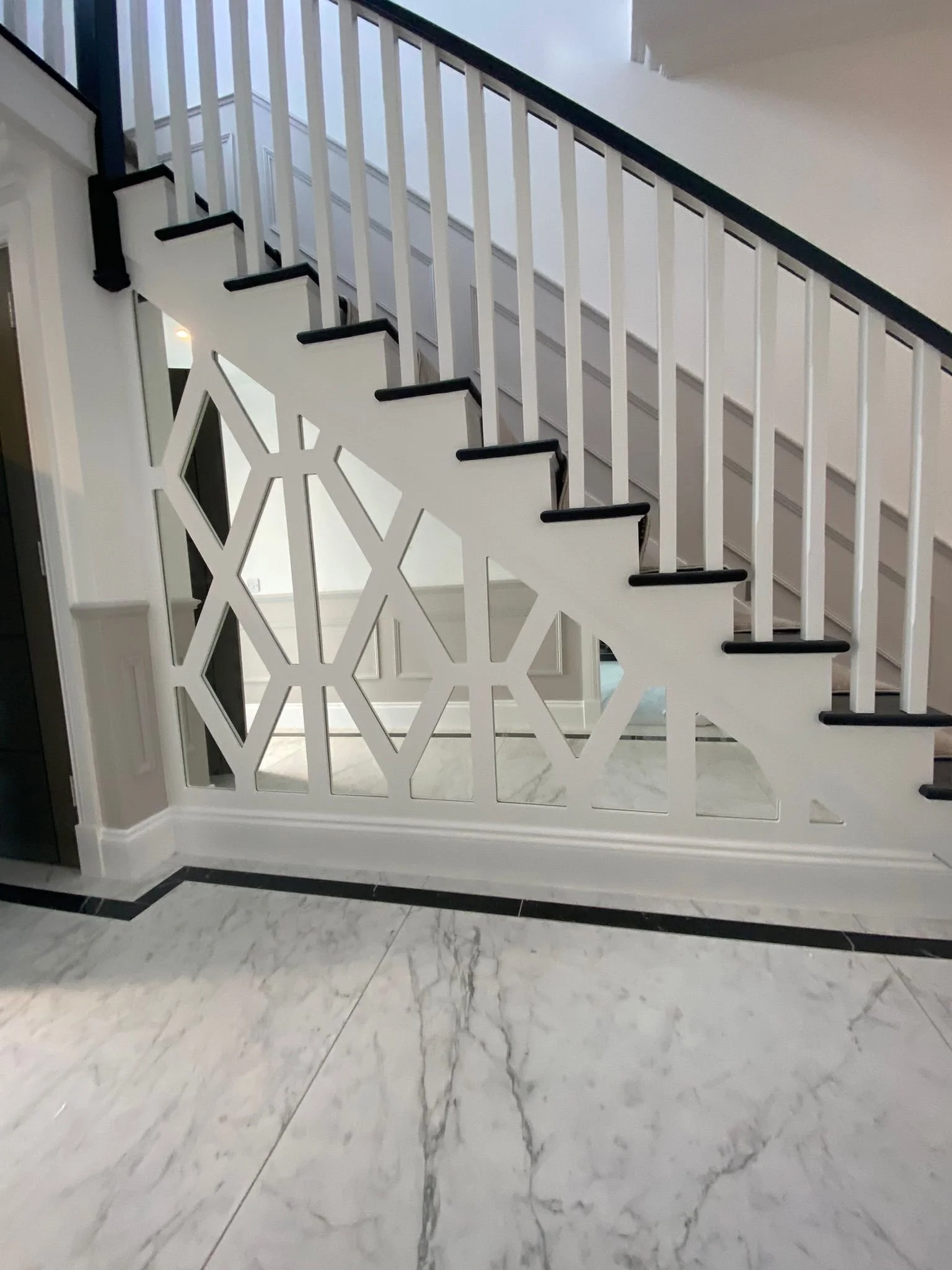 Interior staircase with white railing and black accents, and decorative paneling underneath, inside a home with marble flooring.