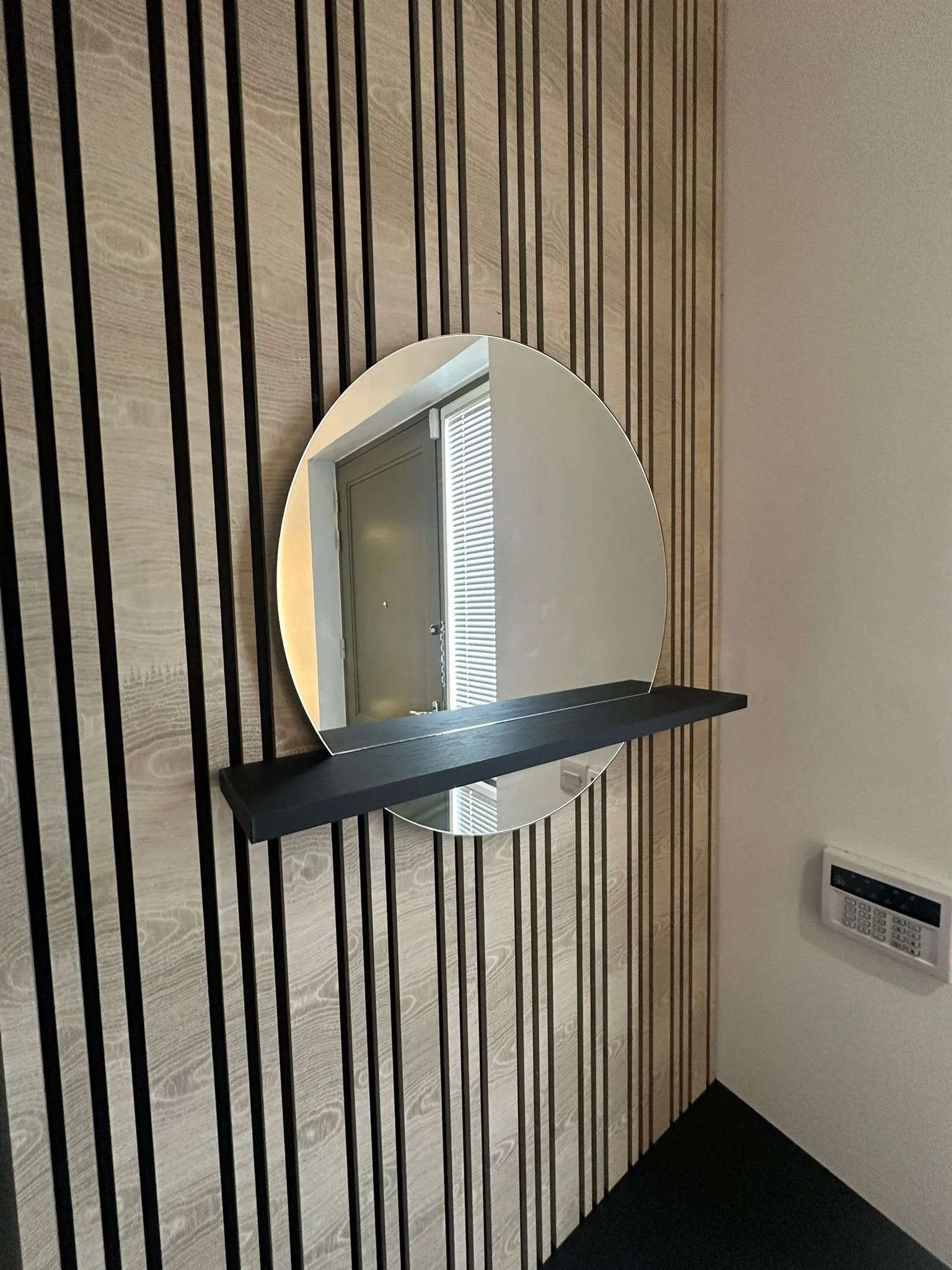 A round mirror on a wall with vertical wooden slats and a black shelf underneath, reflecting a window with blinds and a gray door.