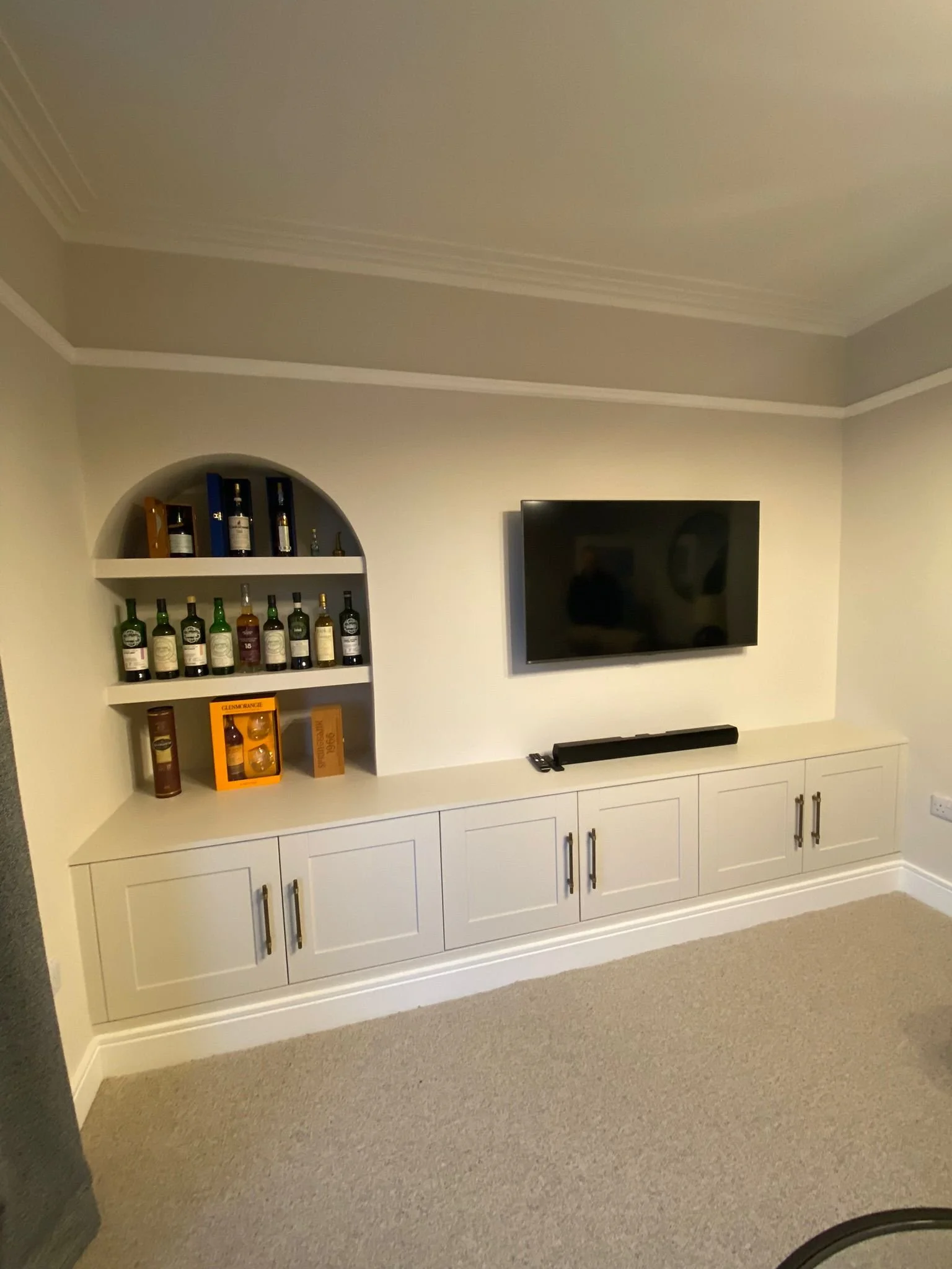 Living room entertainment area with a wall-mounted flat-screen TV, a white cabinet with doors below, a soundbar, and a built-in showcase with bottles of liquor.