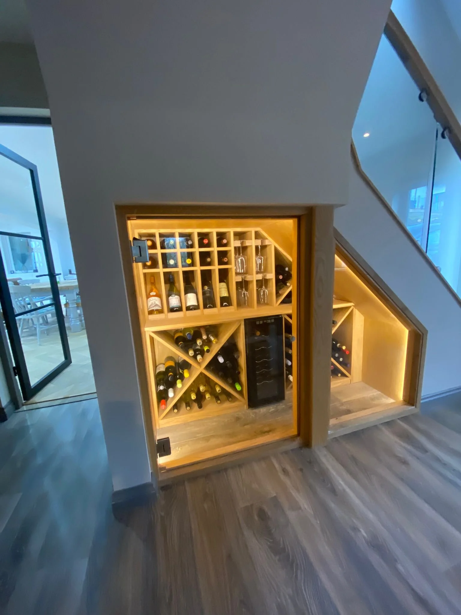Built-in wooden wine storage with bottles and glasses, illuminated by warm lighting inside a modern home near stairs and glass doors.