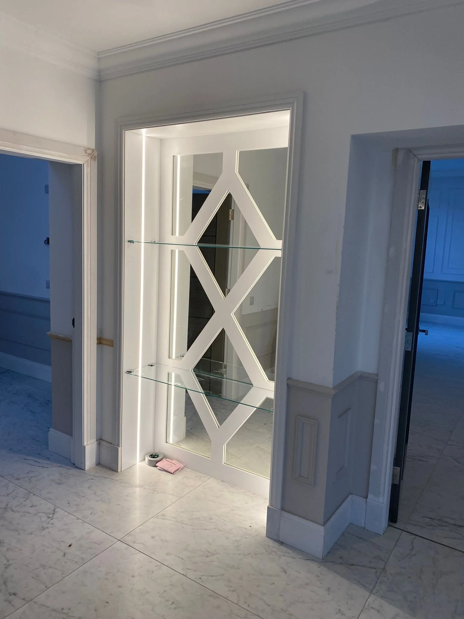 A modern decorative mirror with built-in lighting and glass shelves, framed by white walls and tiled flooring.