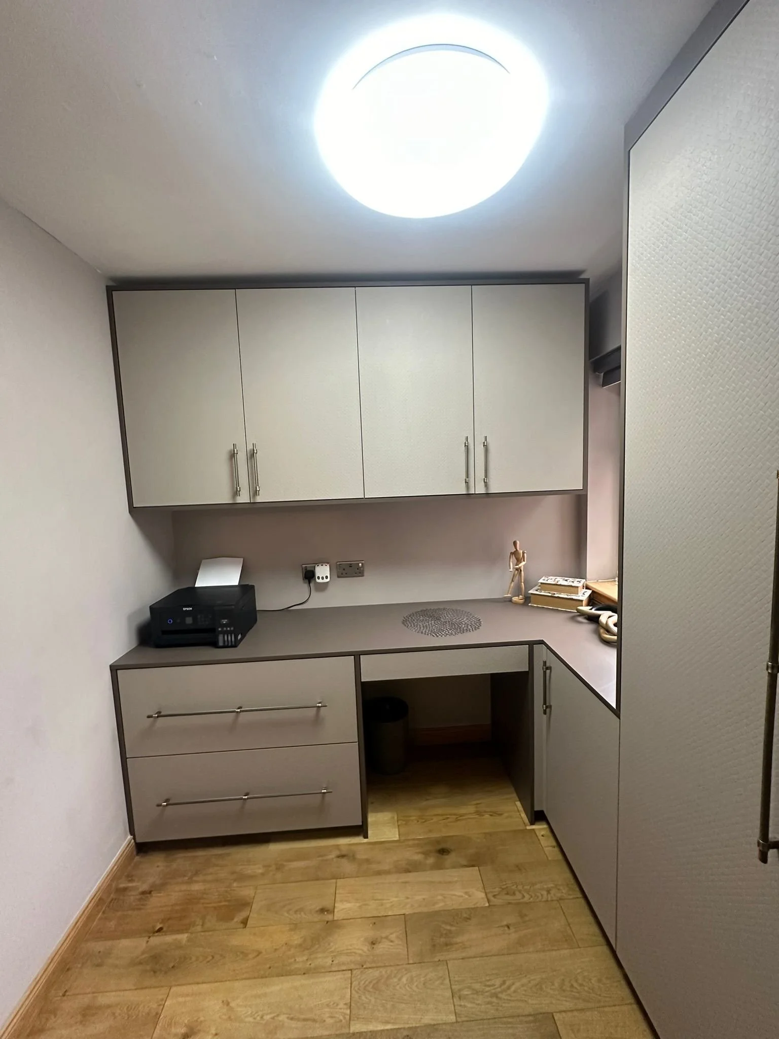 Small office or workspace with upper and lower beige cabinets, a gray countertop, a paper printer, a wooden art mannequin, books, and a window with a shade. The room has wooden flooring and bright ceiling lighting.