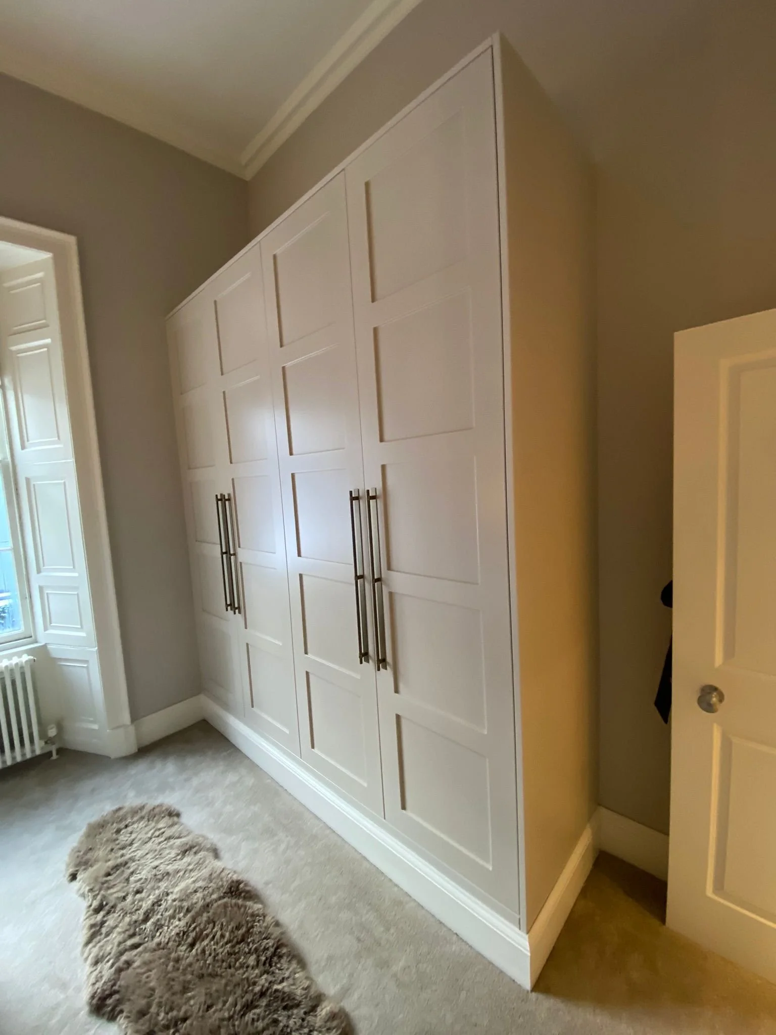 White built-in wardrobe with multiple doors, long handles, in a corner room with beige walls, a window, carpeted floor, and a fluffy rug.