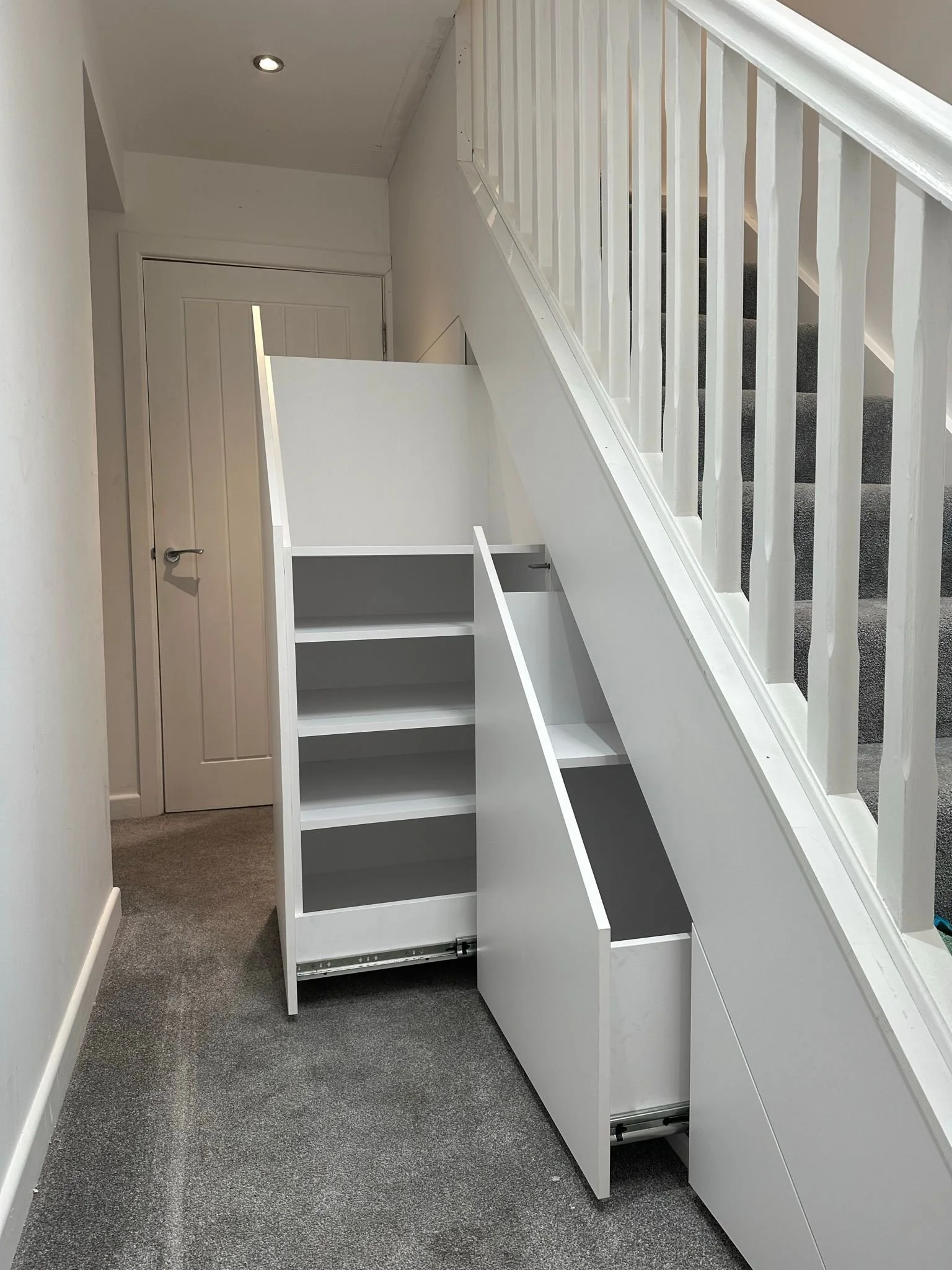 White staircase storage unit with open drawers and shelves, installed under staircase with carpeted stairs and balustrade on the side.