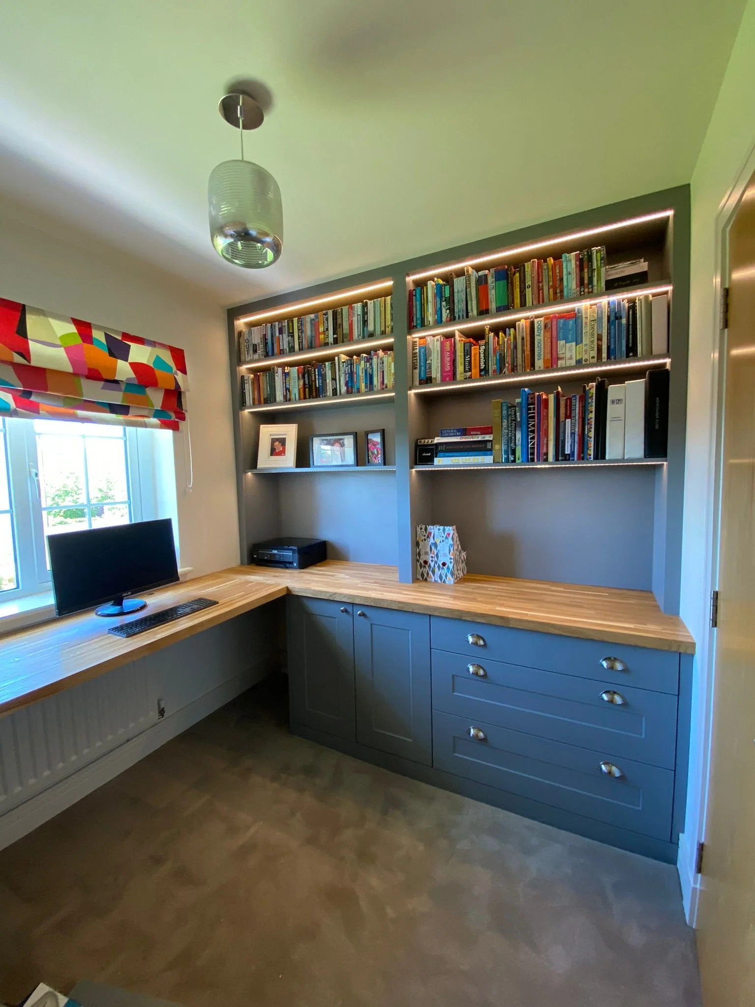 Home office with built-in blue cabinetry, a wooden L-shaped desk, a computer monitor, bookshelves with books and framed photos, window with colorful Roman shade, modern ceiling light, and beige carpet.