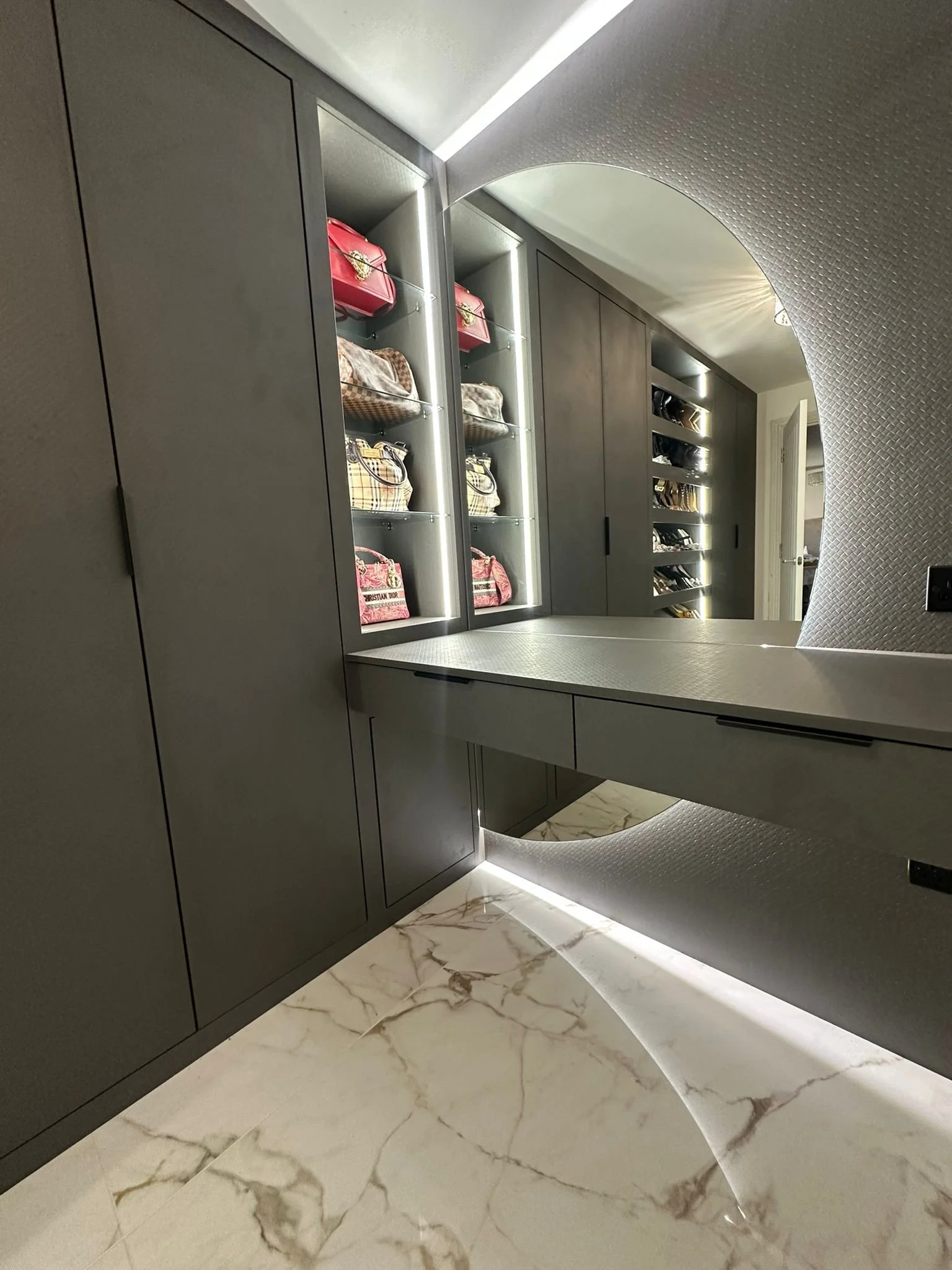 Luxury walk-in wardrobe dressing area with round back-lit mirror and shelves displaying handbags.