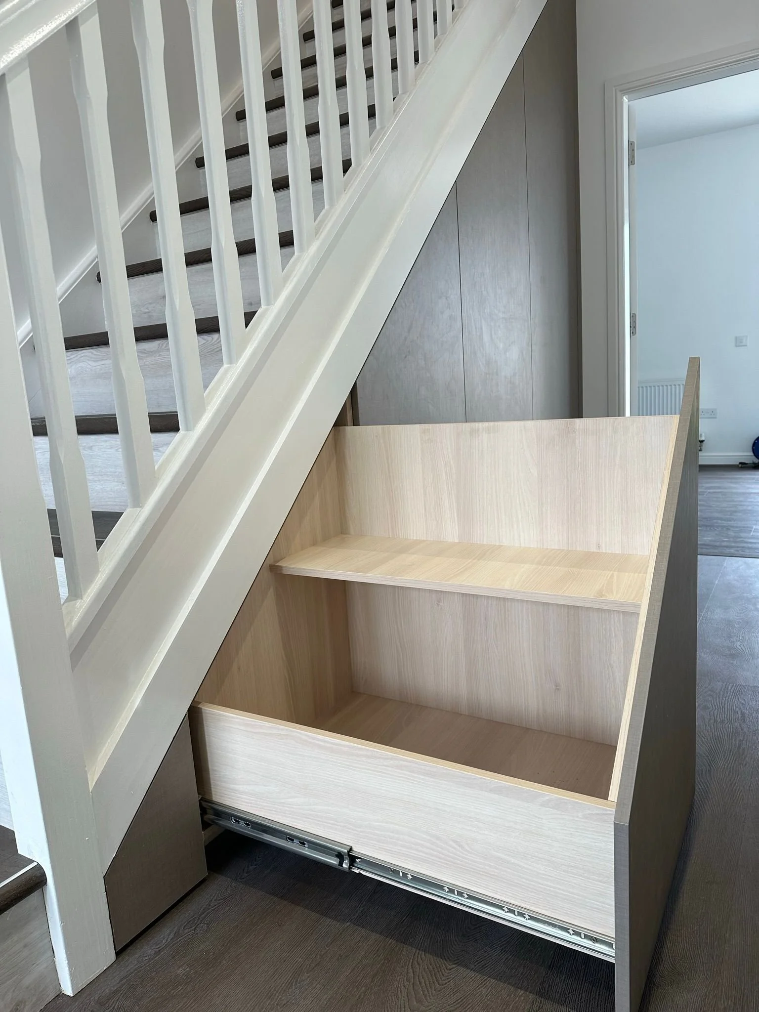Wooden storage unit with a shelf inside, positioned at the bottom of a staircase, with a stair railing above.