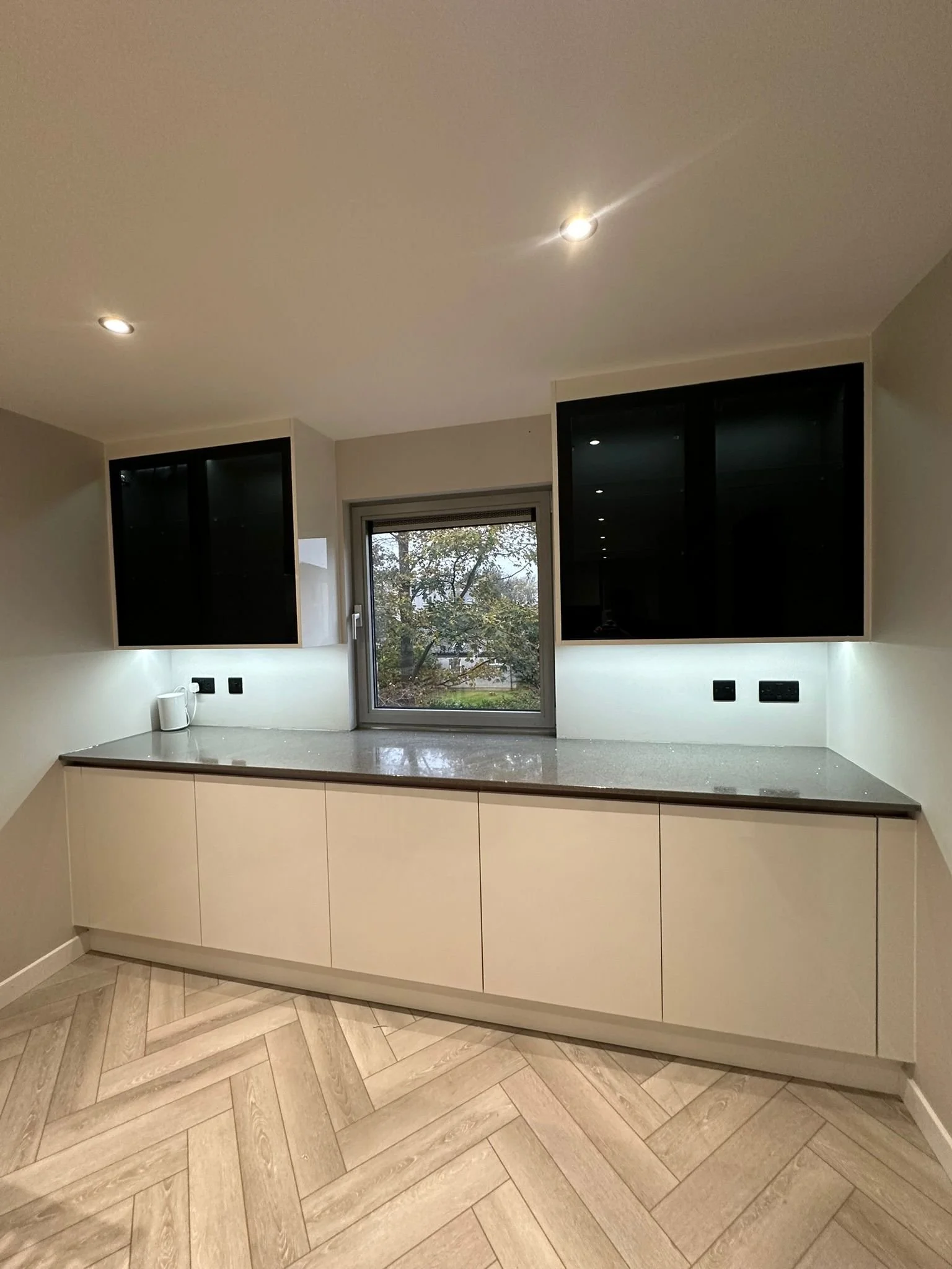 Modern kitchen with grey cabinets, a dark grey countertop, a small window, and two black wall cabinets. Recessed ceiling lights and an outside view of trees visible through the window.