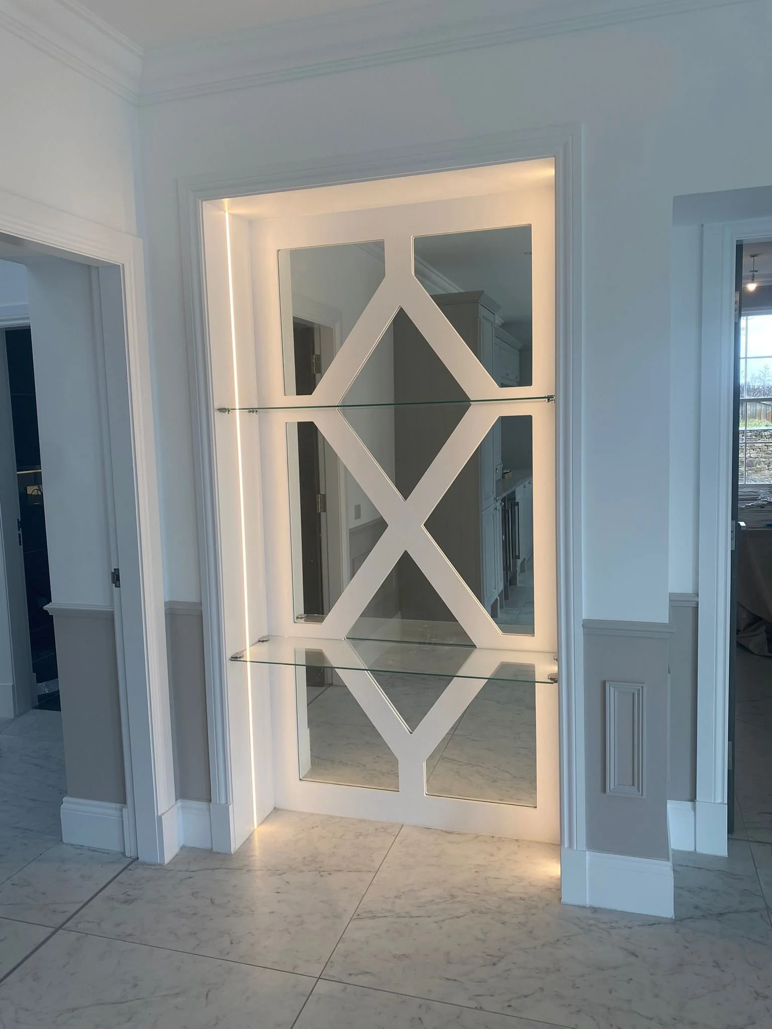 Decorative mirror with geometric white frame and built-in shelves, illuminated by LED lighting, mounted on a white wall in a modern interior.