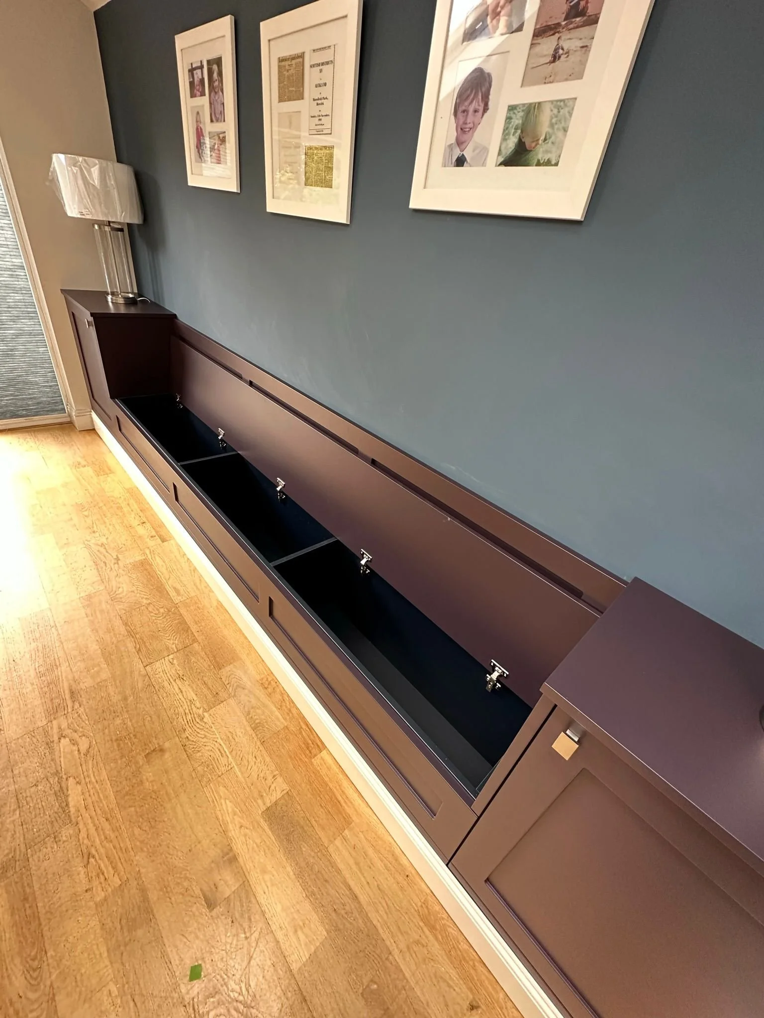 Empty built-in cabinet or media console with open compartments, dark finish, above a light wood floor, on a blue wall with framed pictures.