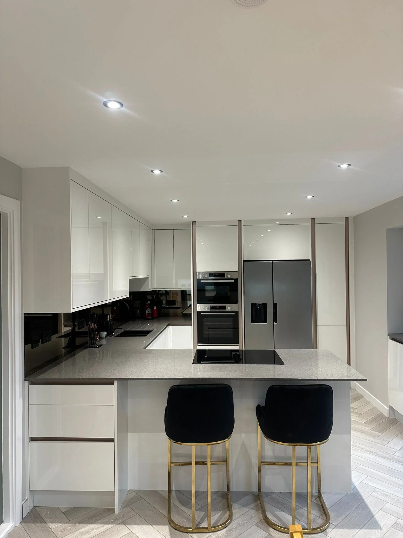 Modern kitchen with white cabinets, dark countertop, stainless steel refrigerator and double oven, two black barstools with gold legs in front of an island, and a light wood floor.