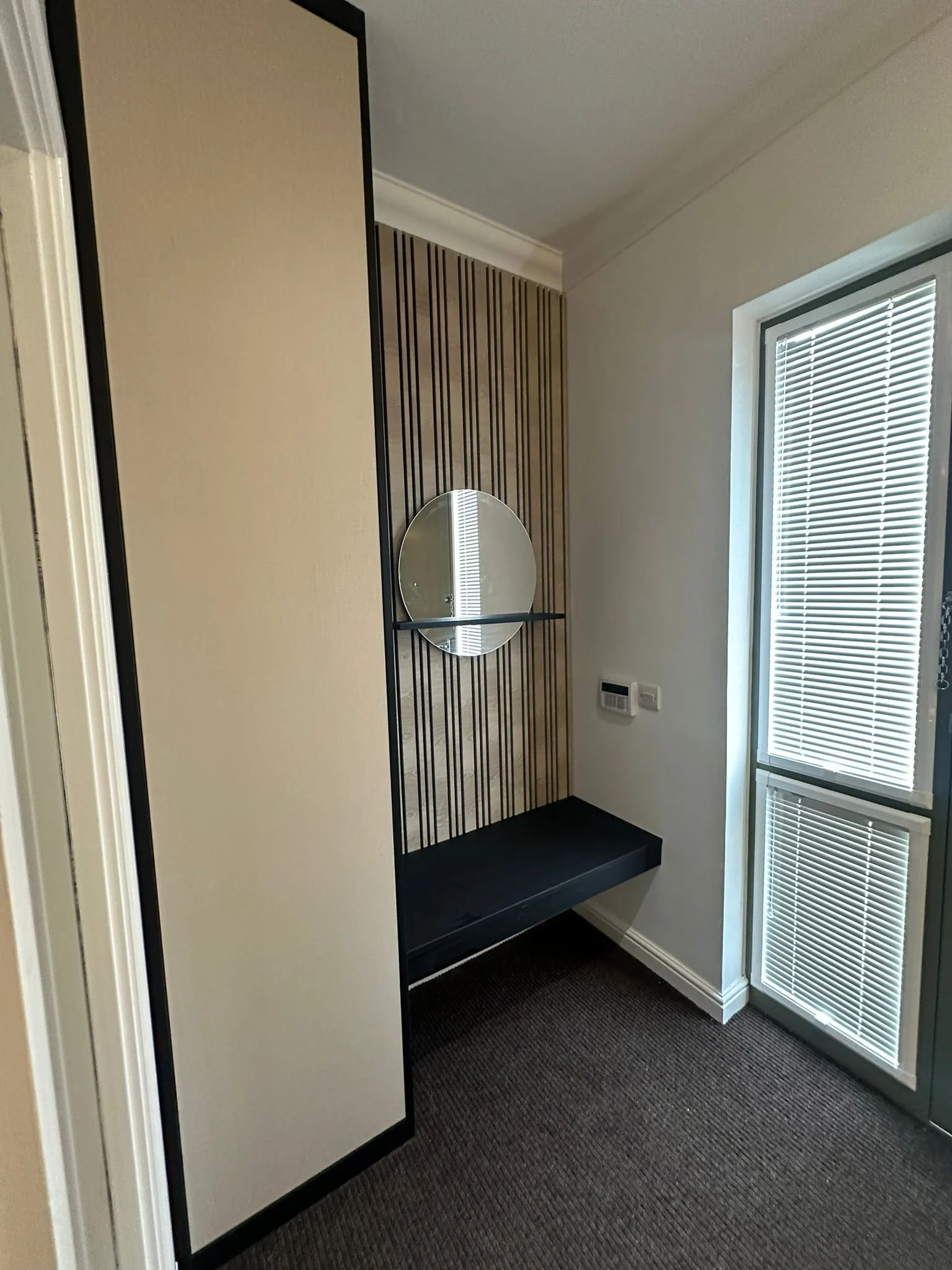 A corner of a room with a mirrored round wall decor, wooden slat panel behind it, a black bench below, a tall cabinet on the left, and a window with closed blinds on the right.