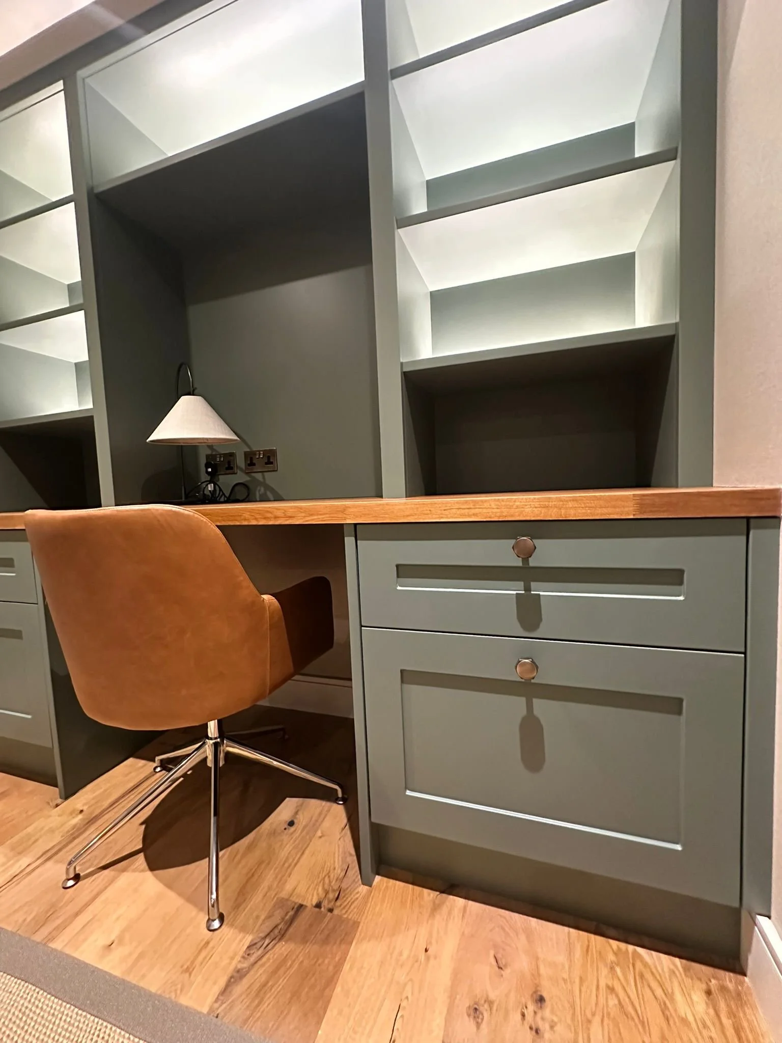 A corner of a room with a green desk and built-in green shelving units above. There is a brown office chair with a chrome base in front of the desk. A small white lamp is on the desk, plugged into an electrical outlet.