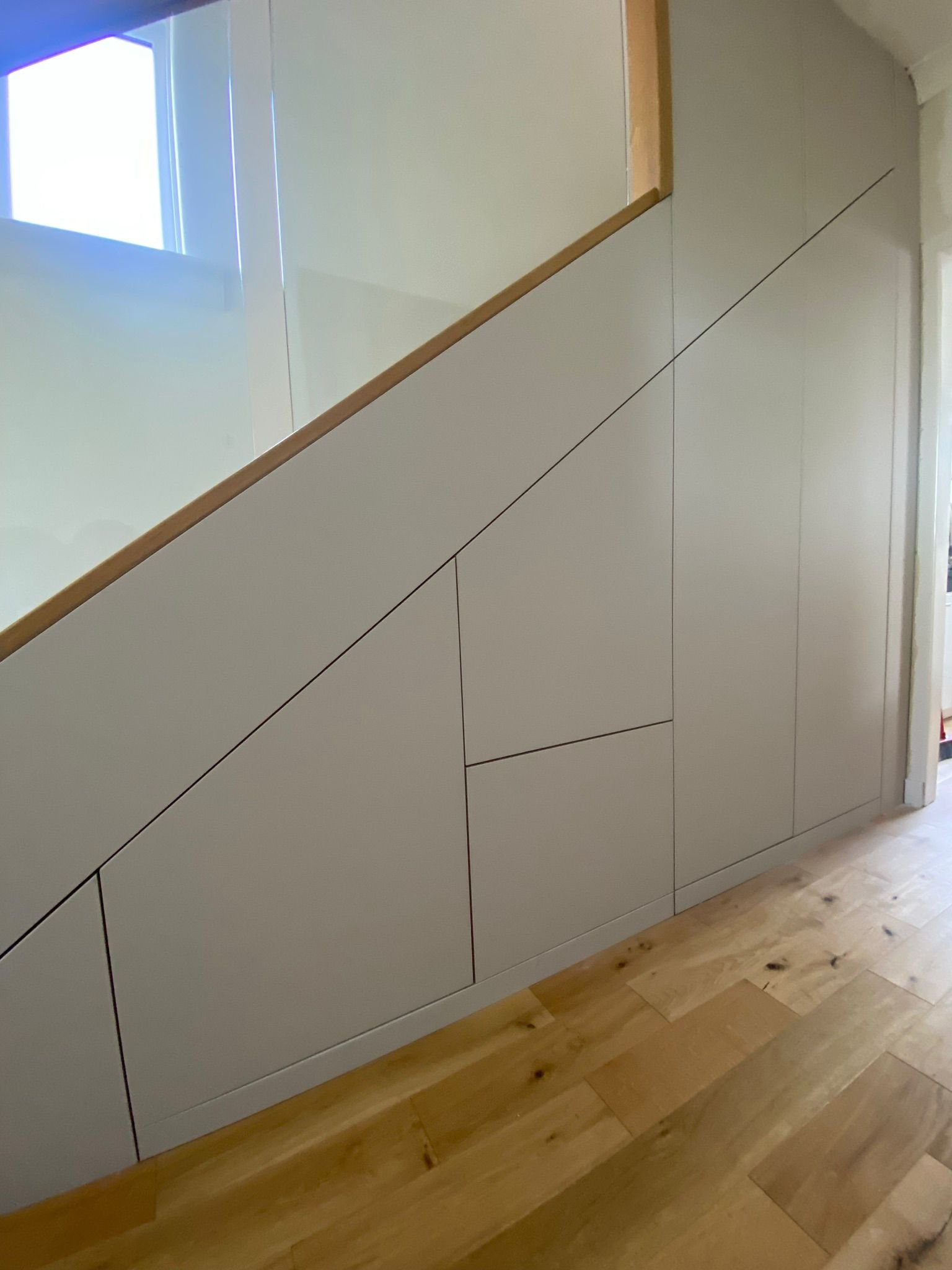 Built-in white cabinets with black outlines beneath a staircase with a wooden handrail, hardwood floor, and a window in the background.