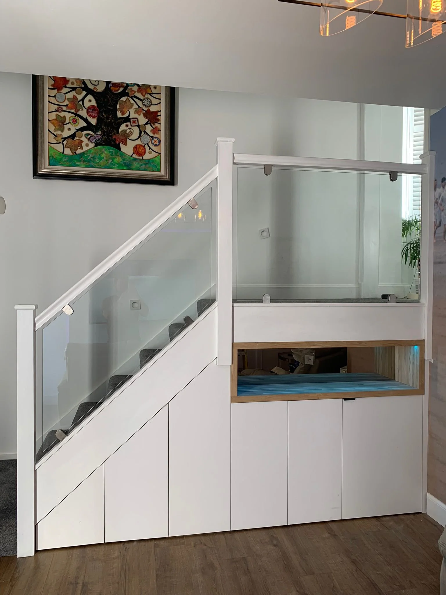 Modern white staircase with glass panels and an open storage area underneath, against a light-colored wall with a colorful framed artwork of a tree.