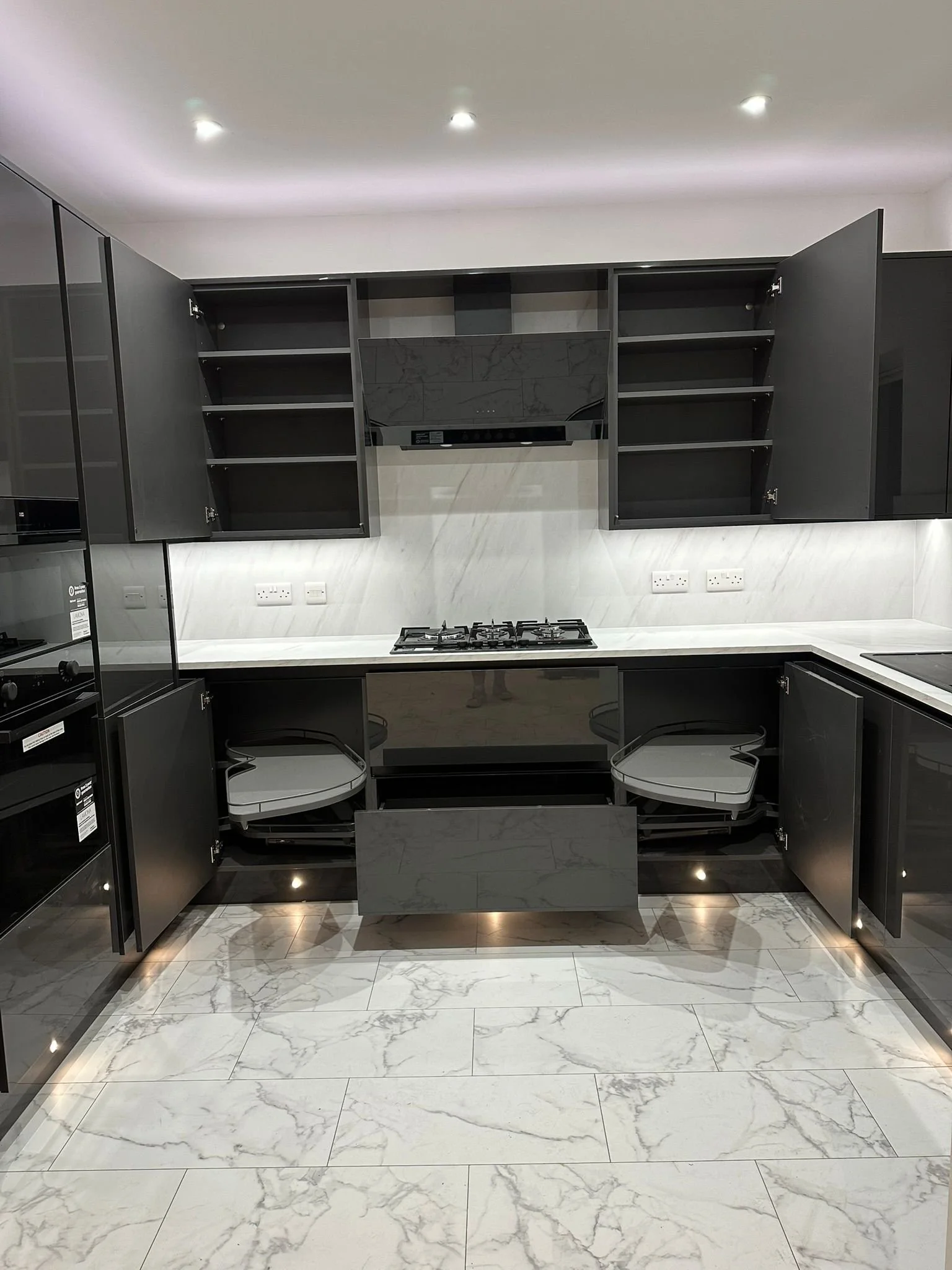 Modern kitchen with black cabinetry, white marble countertops, and built-in appliances including an oven, microwave, and gas stove. The upper cabinets are open, revealing shelves, and the lower cabinets have semi-open pull-out storage.