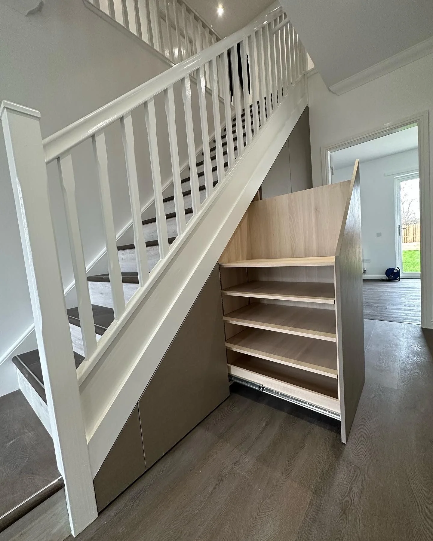 Undermounted wooden shoe storage under staircase in an interior hallway.