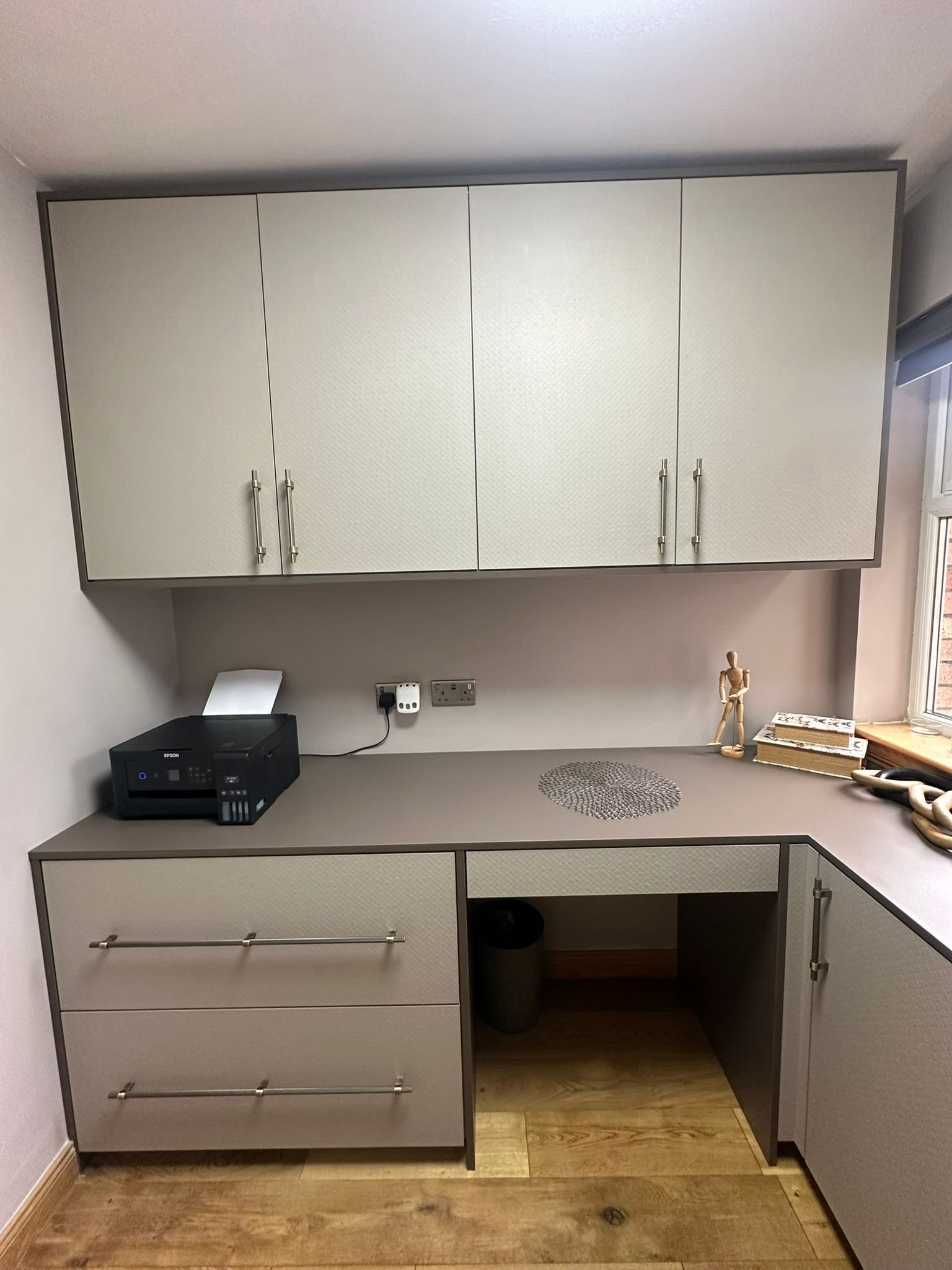 A small built-in office space with light-colored cabinets, a beige countertop, a printer, and a window with natural light.
