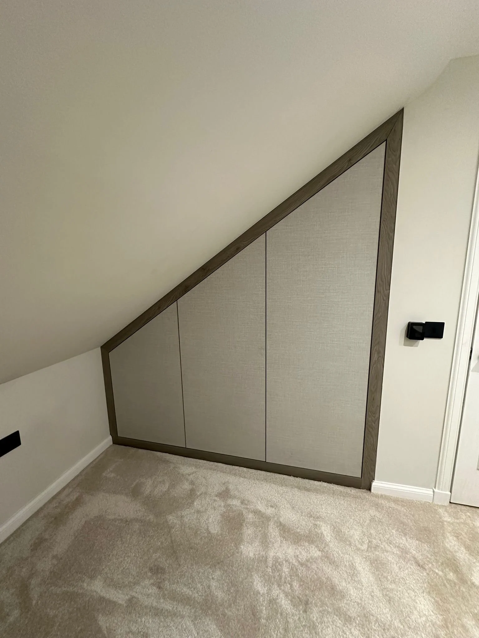 Interior of a room with a sloped ceiling and built-in storage with beige doors and wooden trim, beige carpeted floor, and white walls.
