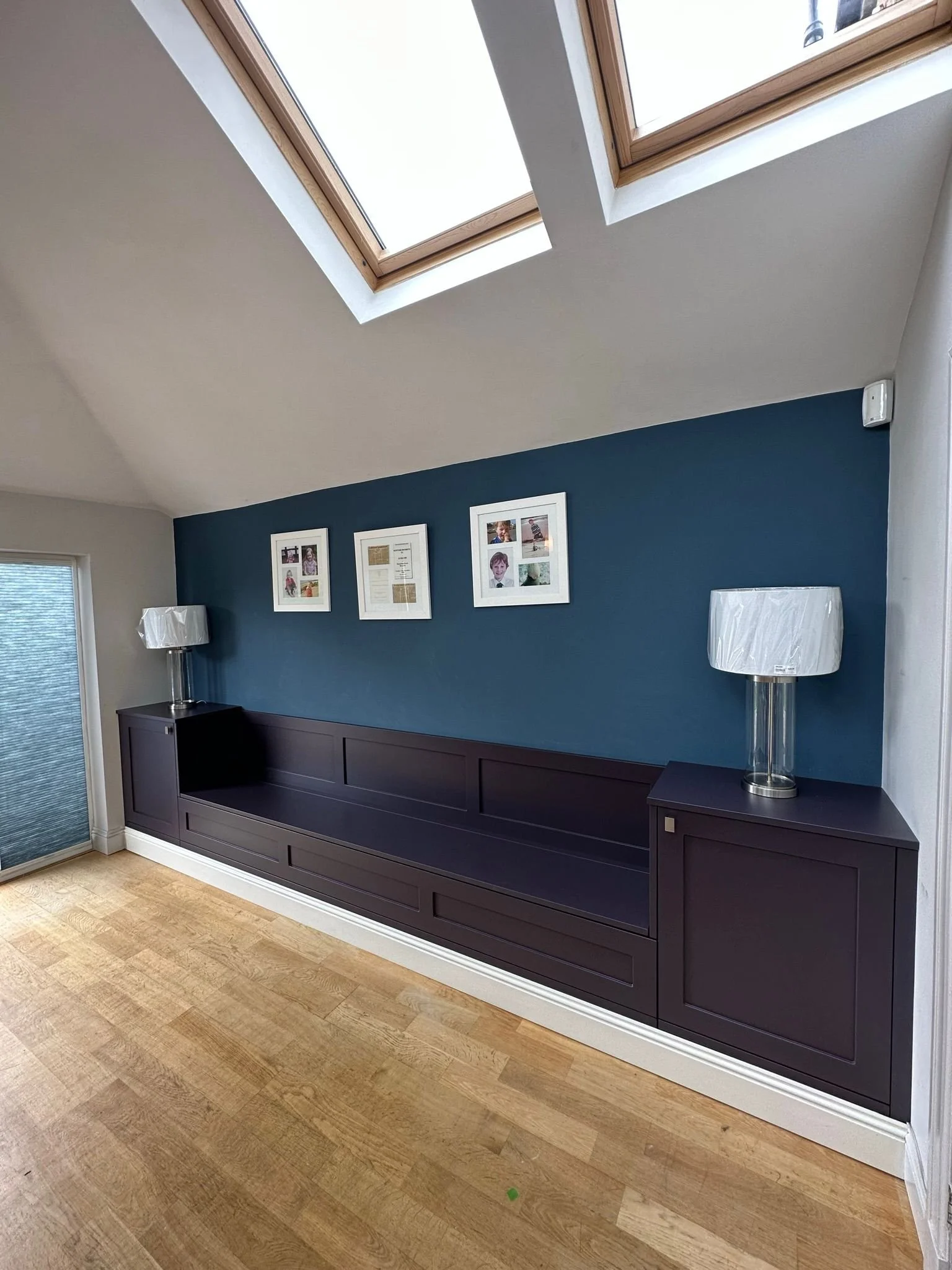 Living room with a dark blue wall, a long built-in dark navy custom built media unit, two glass table lamps, and three framed pictures on the wall. Skylights are visible on the ceiling, and there is a window on the left with blinds.