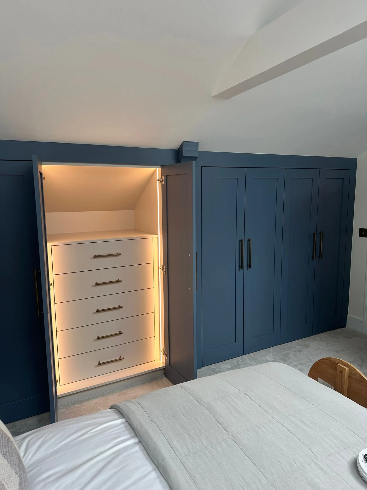 A bedroom with built-in dark blue wardrobes, with one door open revealing a white cabinet with drawers and LED lighting inside. Part of a bed with light-colored bedding is visible at the bottom of the image.