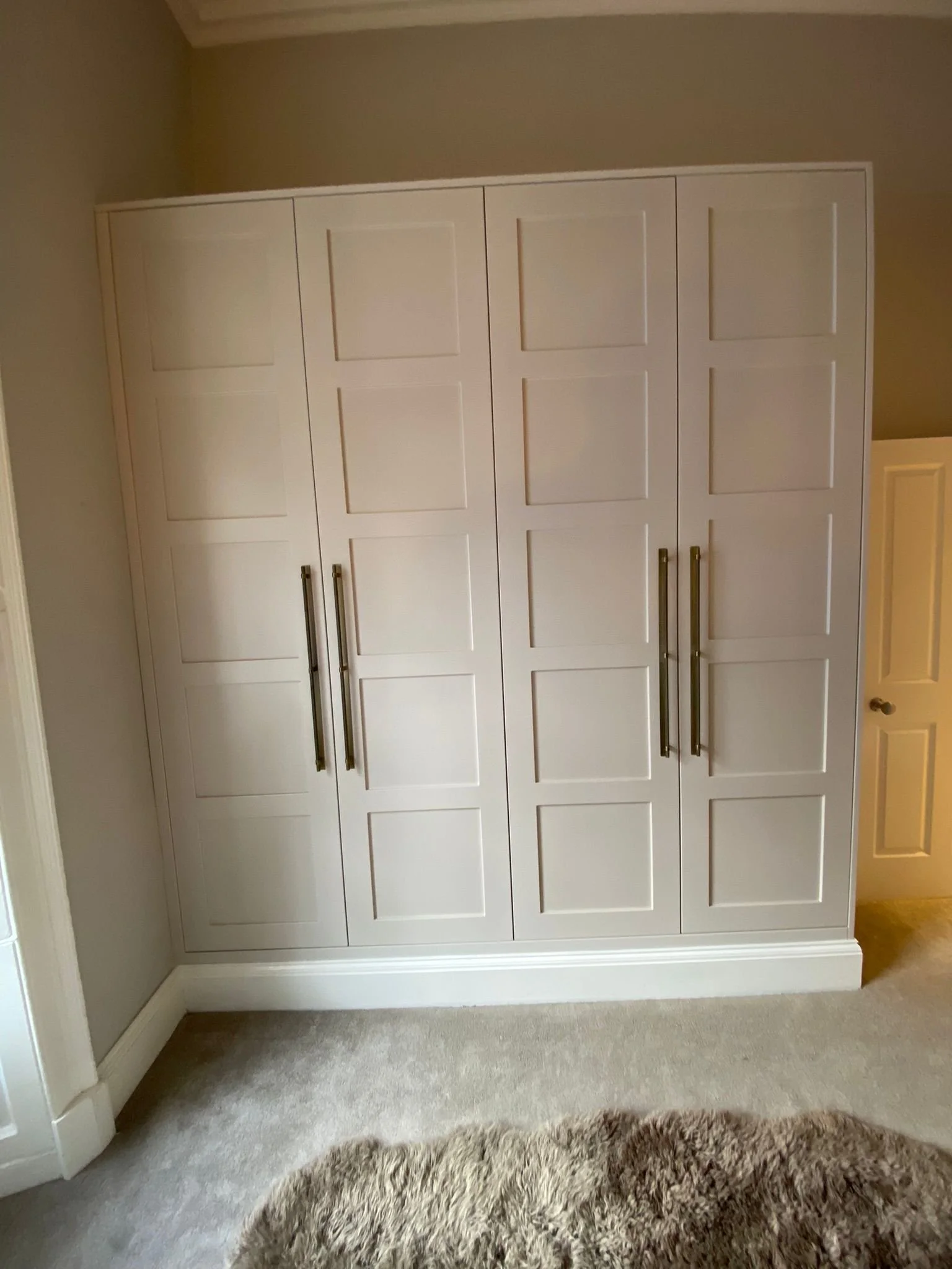 A large white built-in wardrobe with multiple rectangular panels and four vertical brushed nickel handles, situated against a beige wall with beige carpet and a round shaggy beige rug in front.